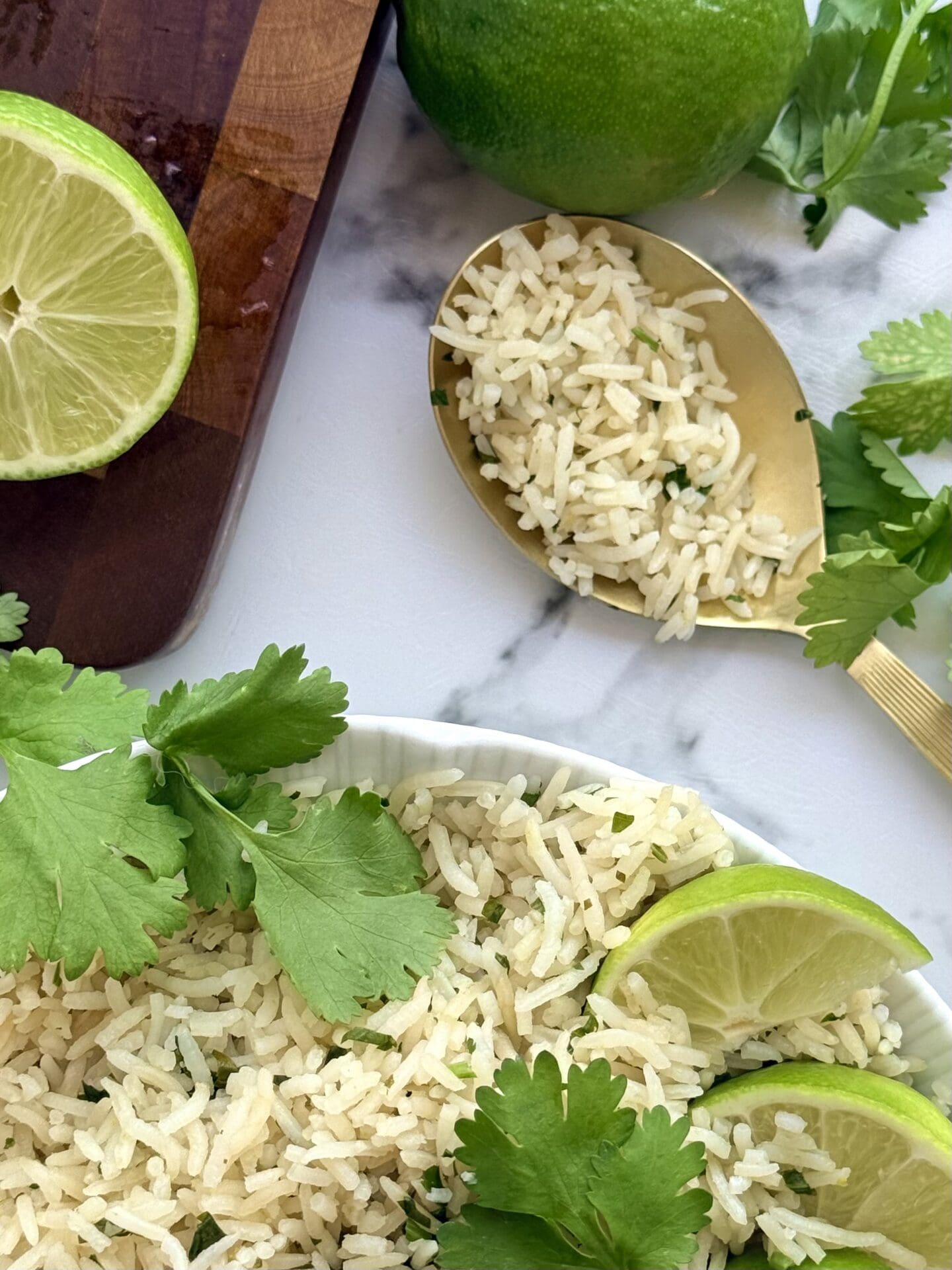IMG_8973 A gold coloured serving spoon is filled with a scoop of cilantro lime rice. The spoon rests on a white marble counter near a dark wood cutting board. Fresh lime and cilantro complete the scene.