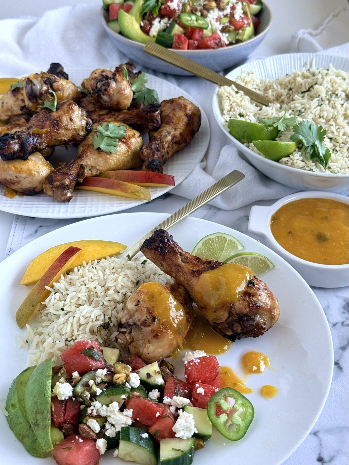 IMG_9039 A dinner plate is seen in the foreground, filled with a serving of vibrant watermelon avocado salad, mango glazed chicken drumsticks and cilantro lime rice. Full platter of each of these dishes is seen in the background.