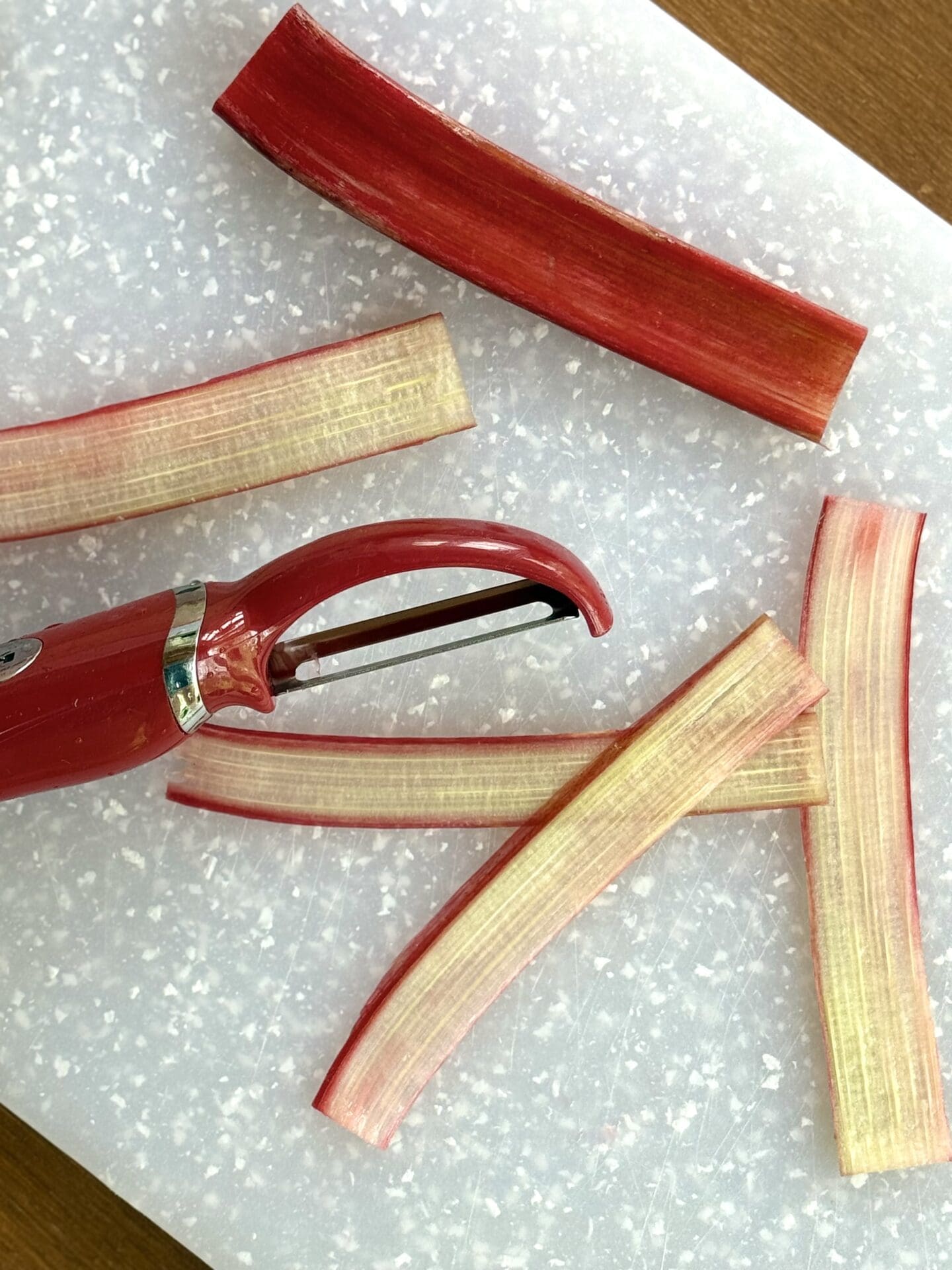 A demonstration photo showing how a vegetable peeler is used to cut fresh rhubarb into long, thin strips to make candied rhubarb ribbons.