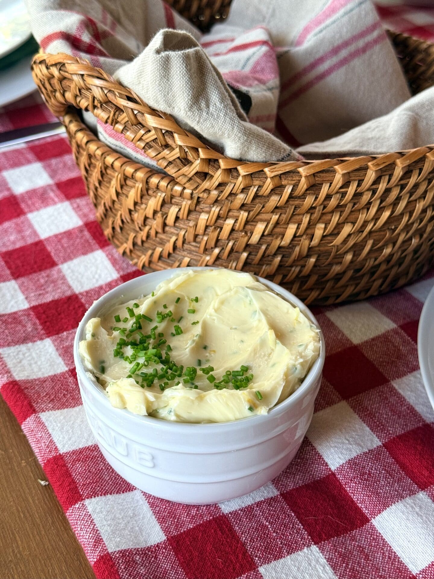 A small bowl of Lemon Chive Butter sits on a red and white gingham tablecloth, near a basket of fresh bread. The top of the butter is generously garnished with chopped fresh chives.