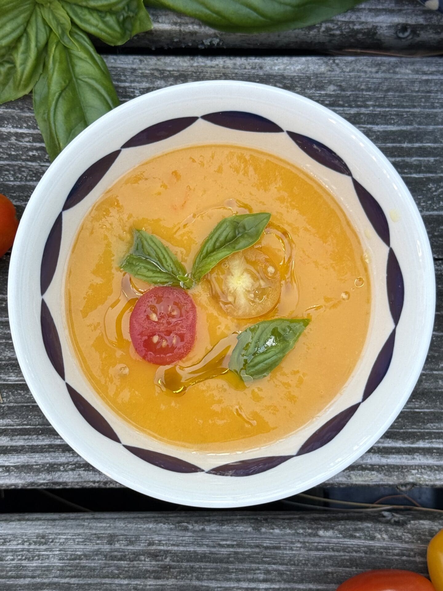 IMG_9809 A single bowl of bright Yellow Tomato Gazpacho is seen from above on a wood table. The soup is garnished with cherry tomatoes and fresh basil.