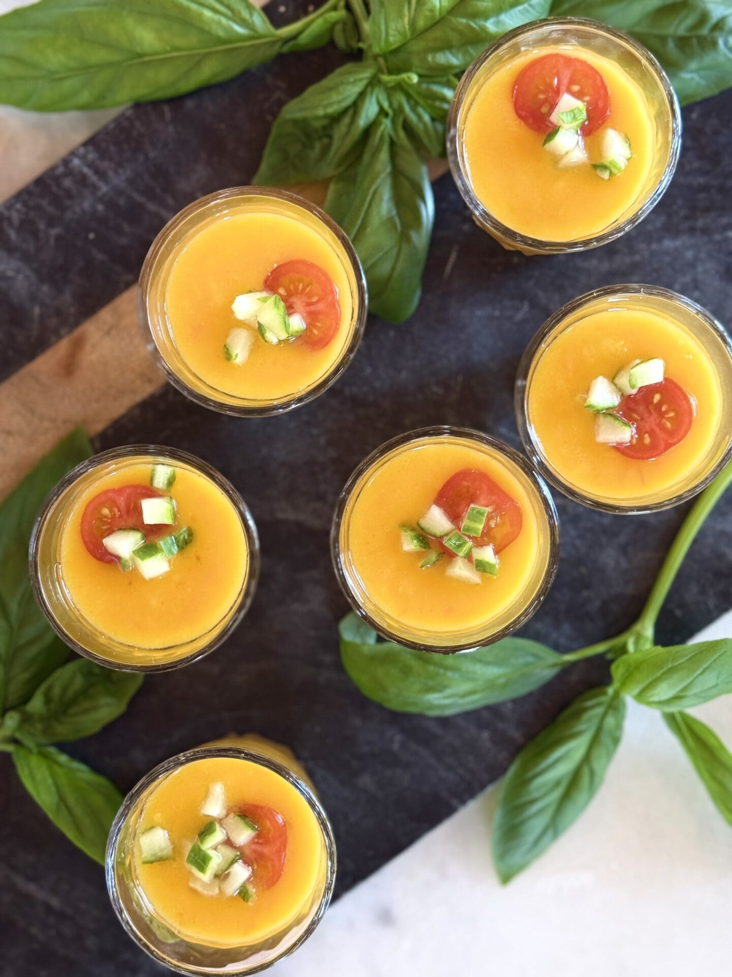 IMG_9832 Small shot glasses filled with Yellow Tomato Gazpacho are seen from above. The soup is garnished with fresh cucumber and cherry tomato slices, and surrounded by fresh basil leaves.