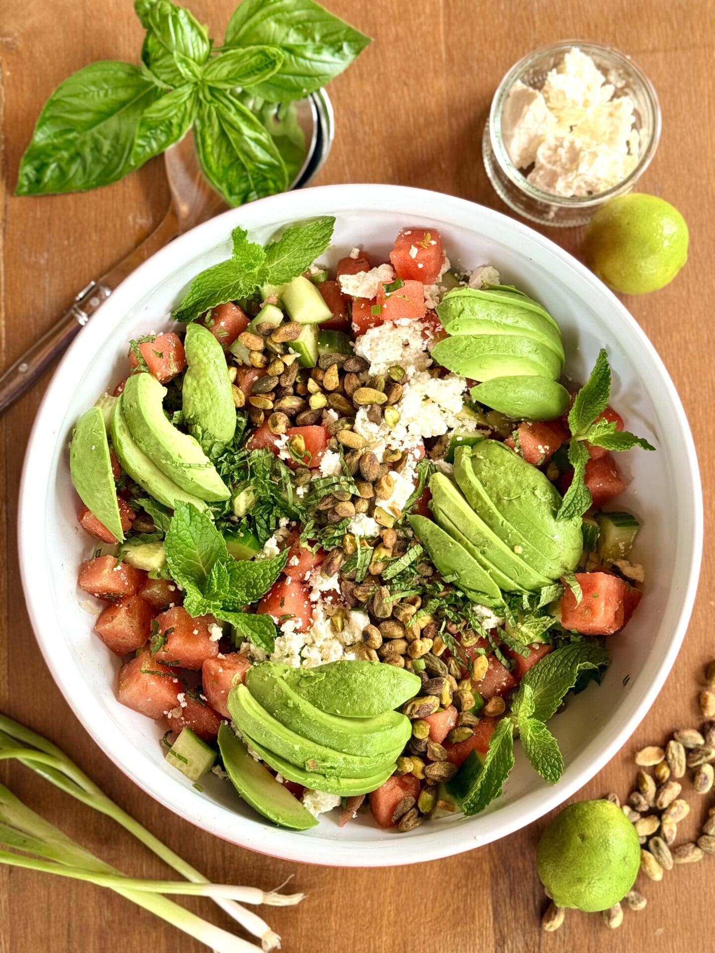 A large bowl of watermelon feta salad with avocado, pistachios and mint is seen from above, surrounded by fresh vegetables, fruit and herbs.