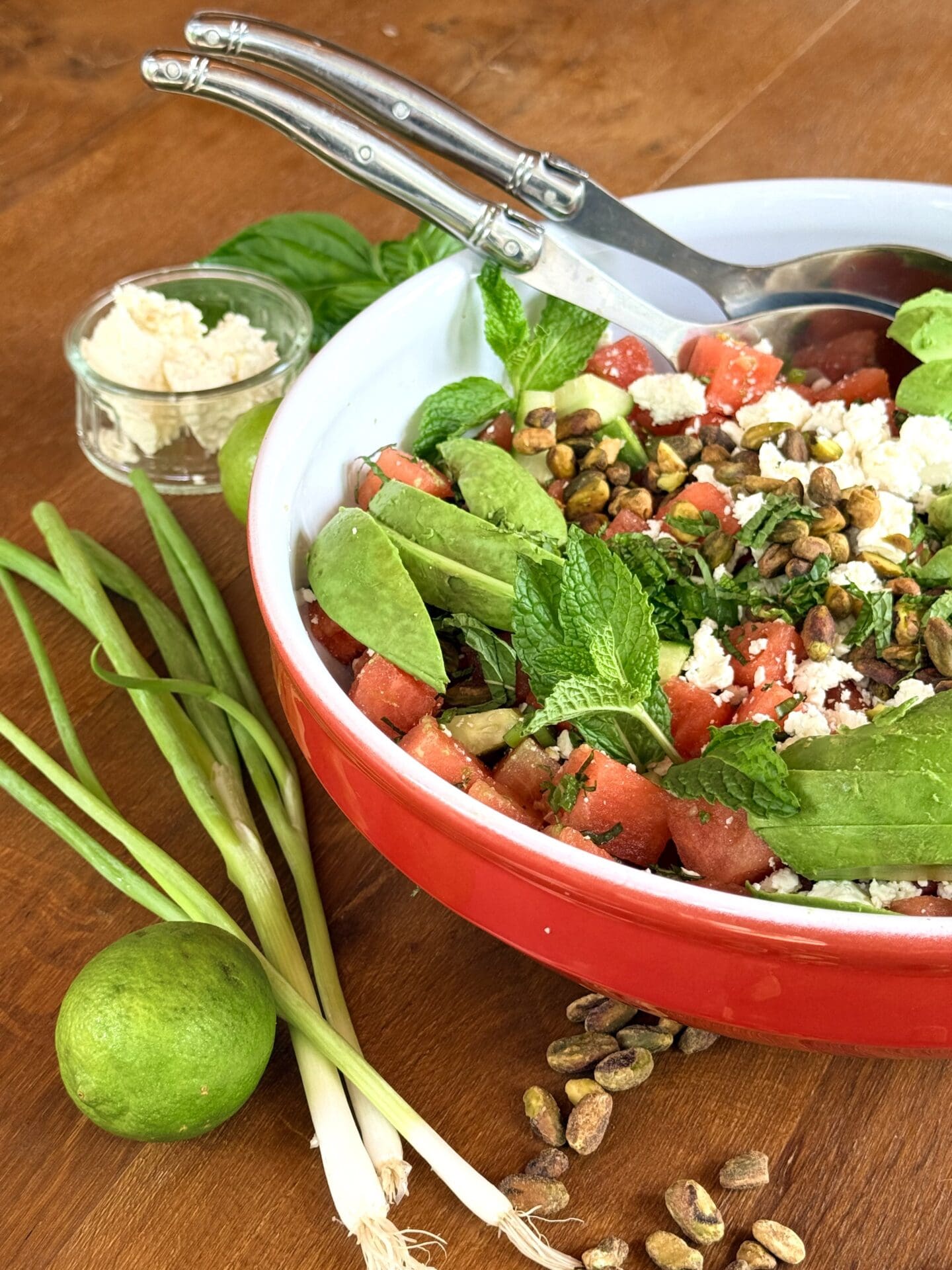 IMG_0262 A large red bowl filled with watermelon feta salad sits on a wood table, surrounded by feta cheese, limes, green onions and pistachios.
