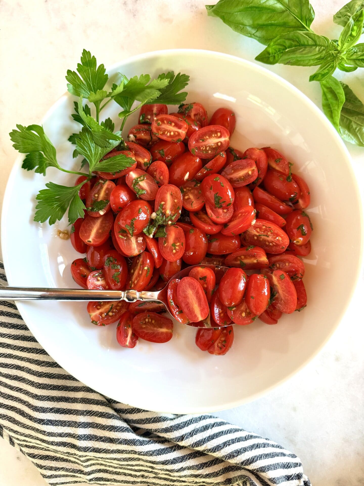 A bowl of marinated cherry tomatoes is seen from above, surrounded by fresh herbs.