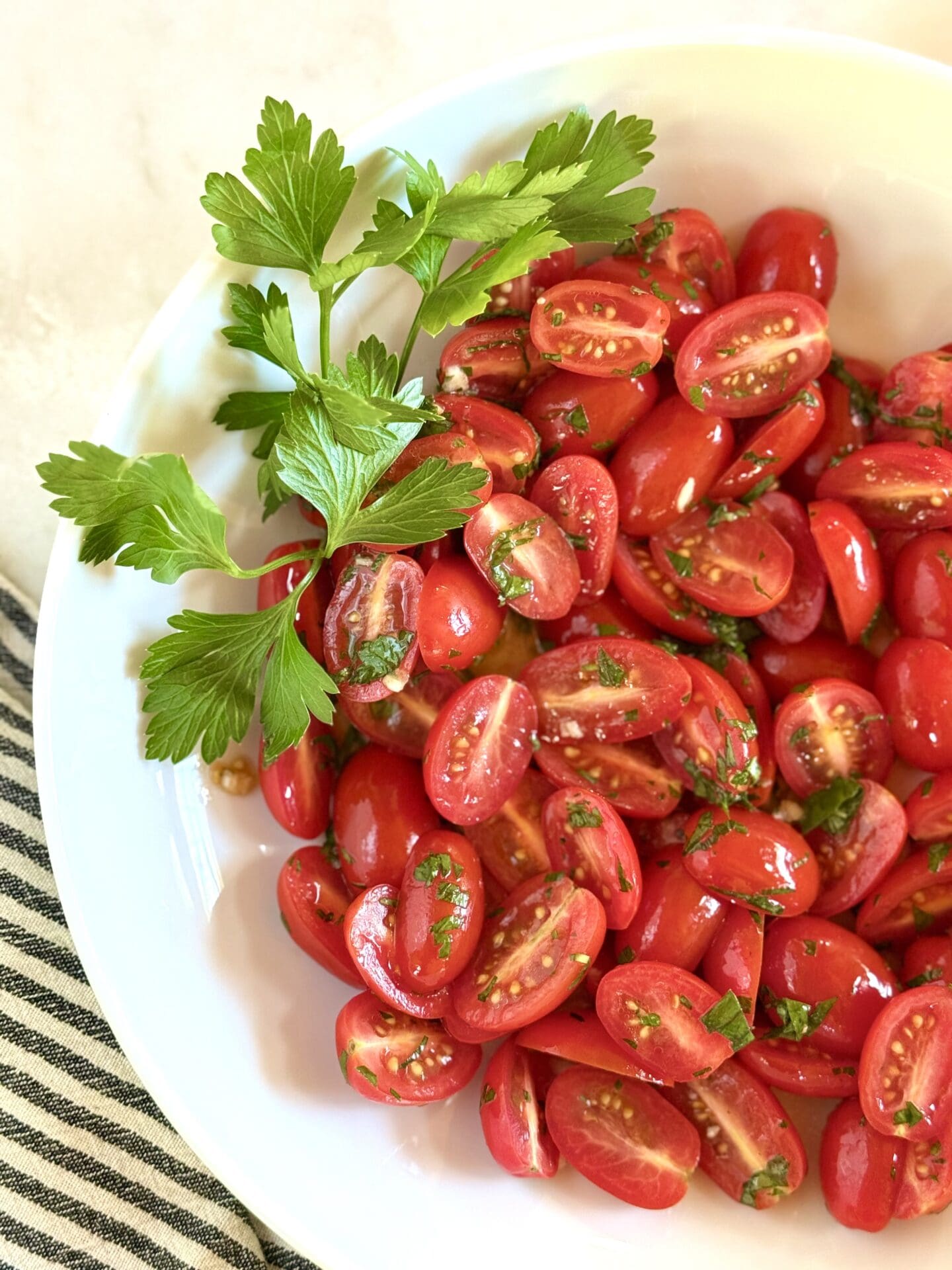 A bowl of marinated cherry tomatoes sits on a white marble countertop, surrounded by fresh herbs.
