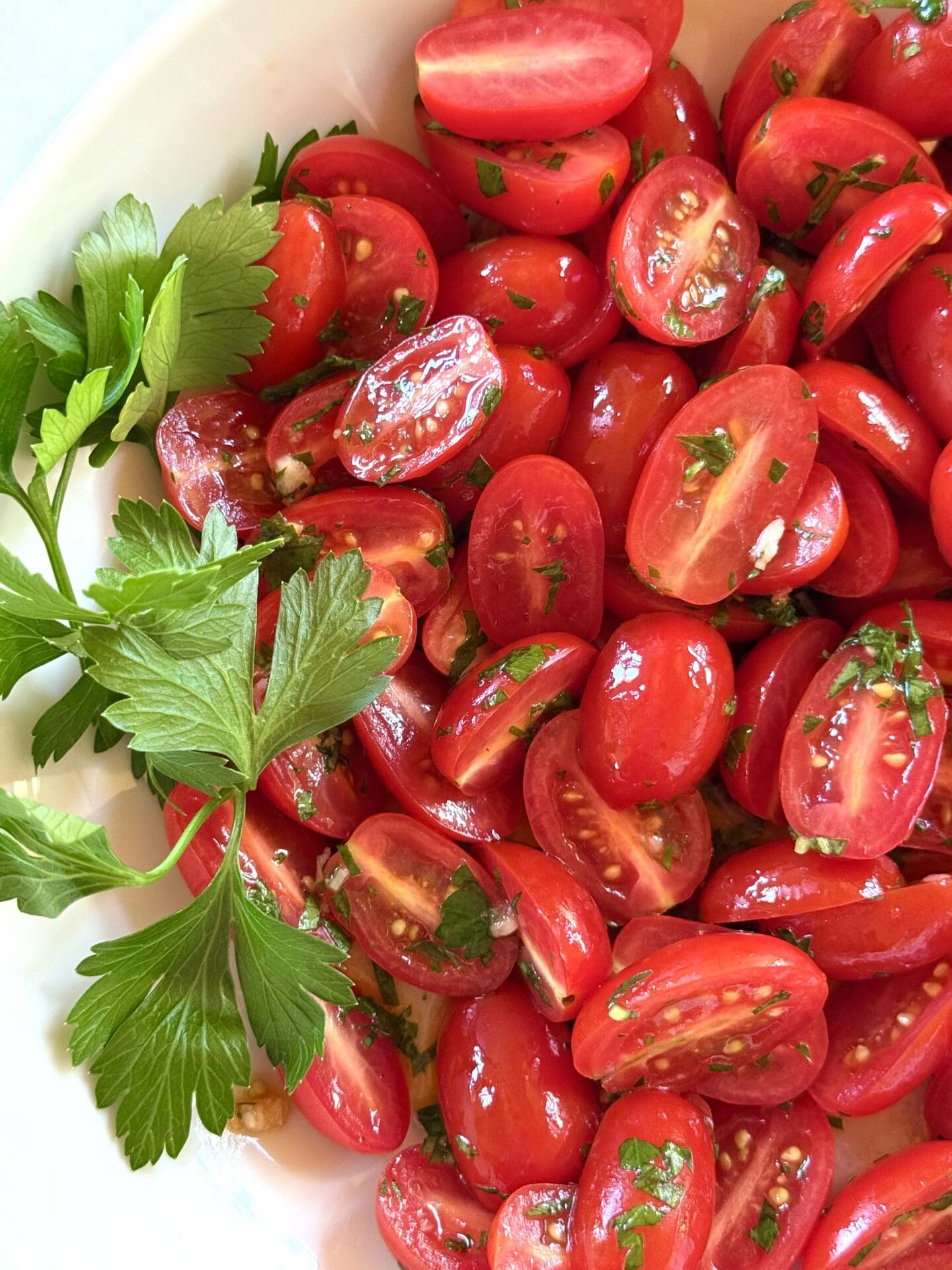 A bowl of marinated cherry tomatoes is seen from above and close up. The image shows the juicy interiors of the tomatoes, flecked with fresh herbs.