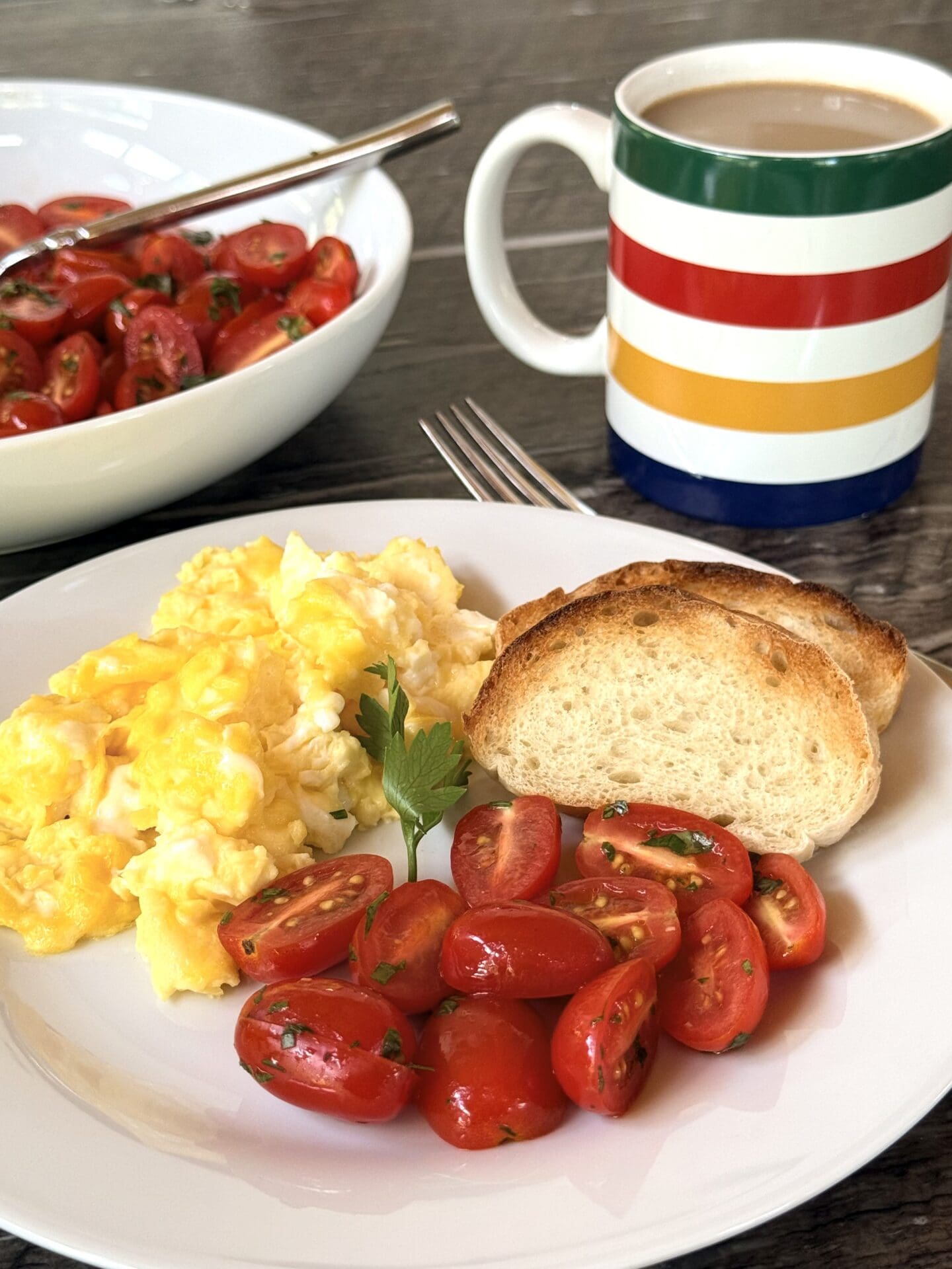 A breakfast plate filled with scrambled eggs, toasted baguette and marinated cherry tomatoes is seen in the foreground. In the background a serving bowl full of the tomatoes and a Hudson Bay Company striped mug of coffee are seen.