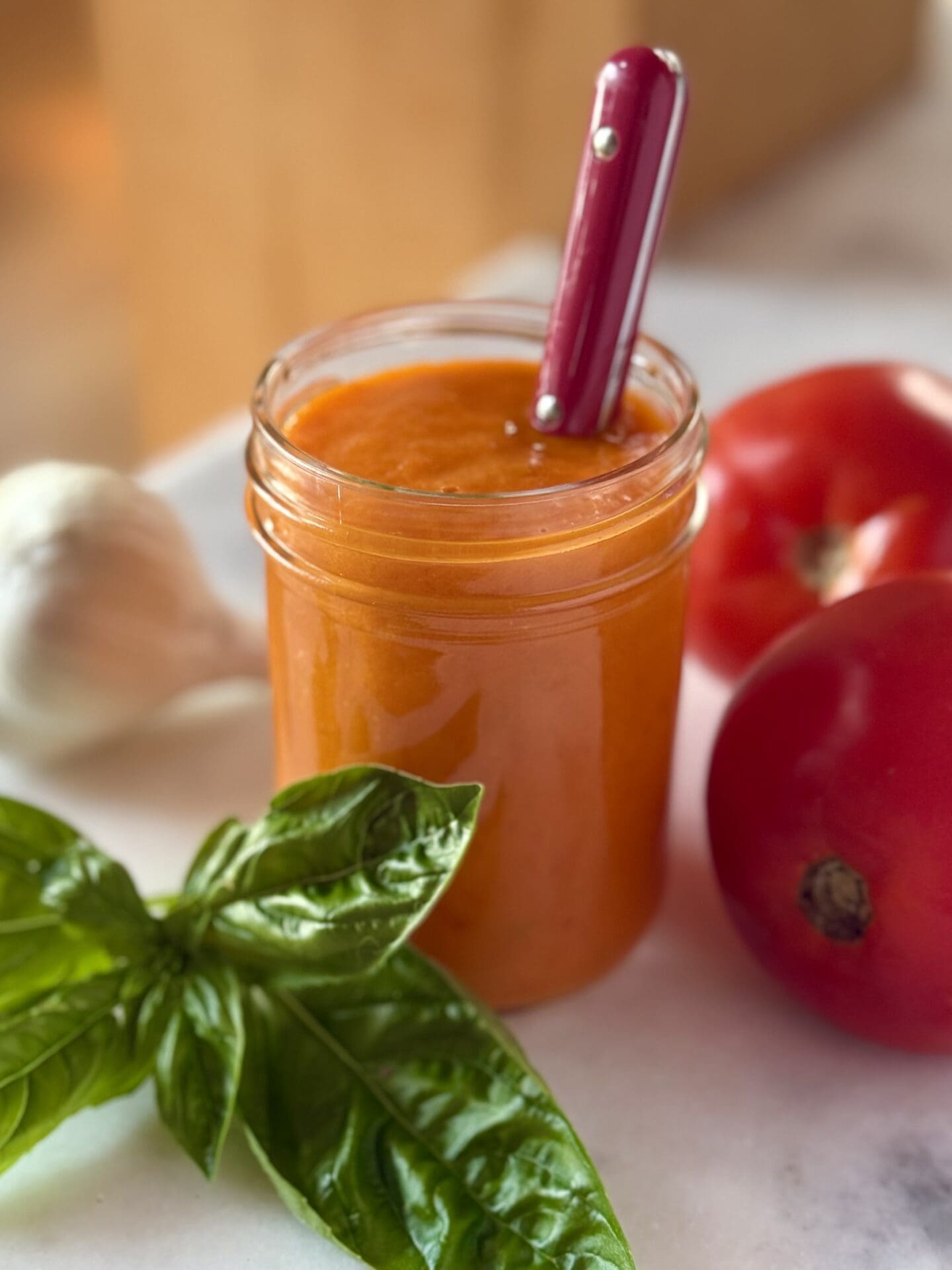 A glass jar filled with fresh tomato sauce is seen sitting on a white marble counter. Fresh tomatoes, basil and garlic surround the sauce.