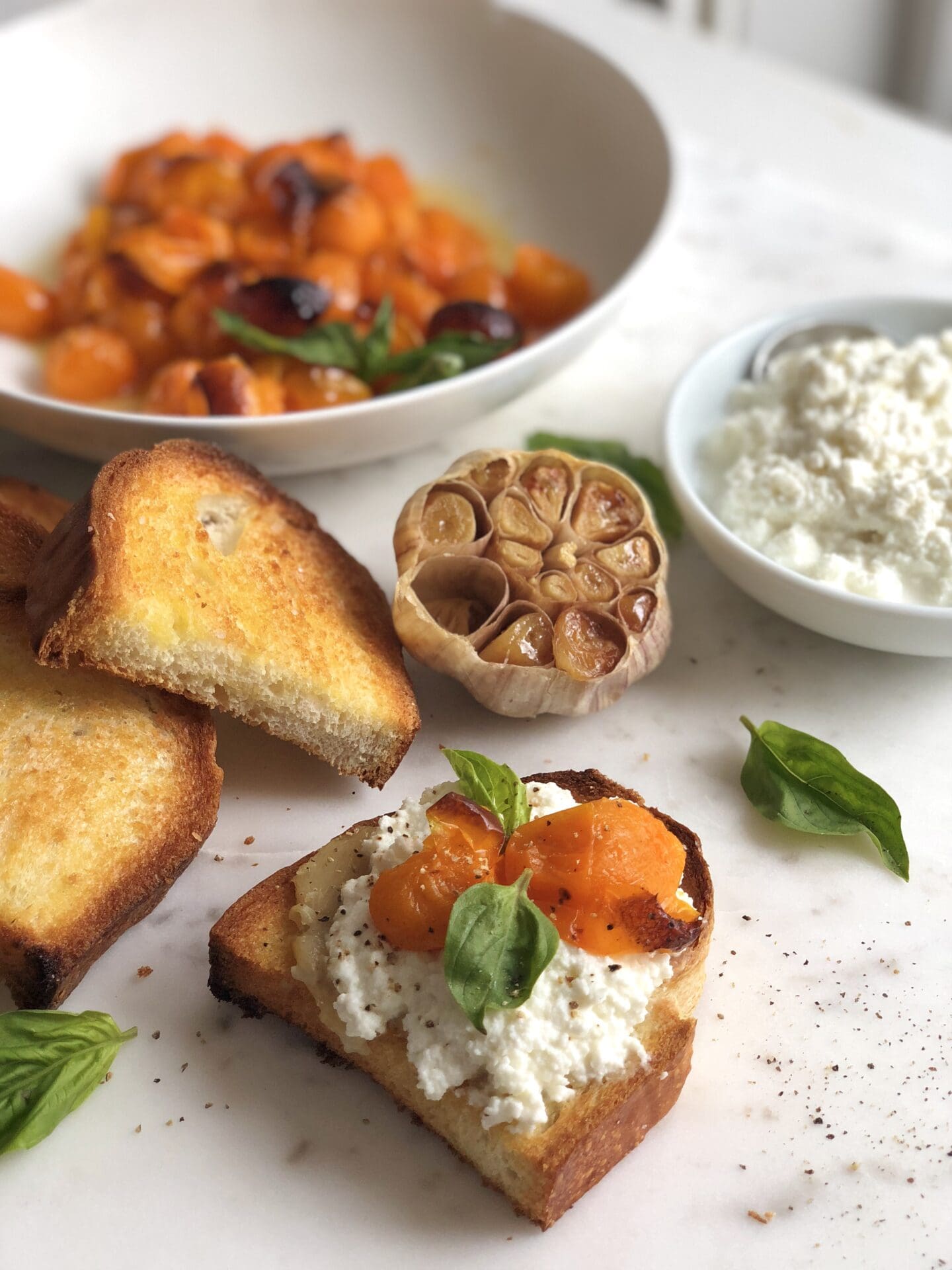 Crostini is made from a larger slice of Italian bread and spread with fresh ricotta and roasted yellow tomatoes. Roasted garlic and fresh basil leaves complete the image.
