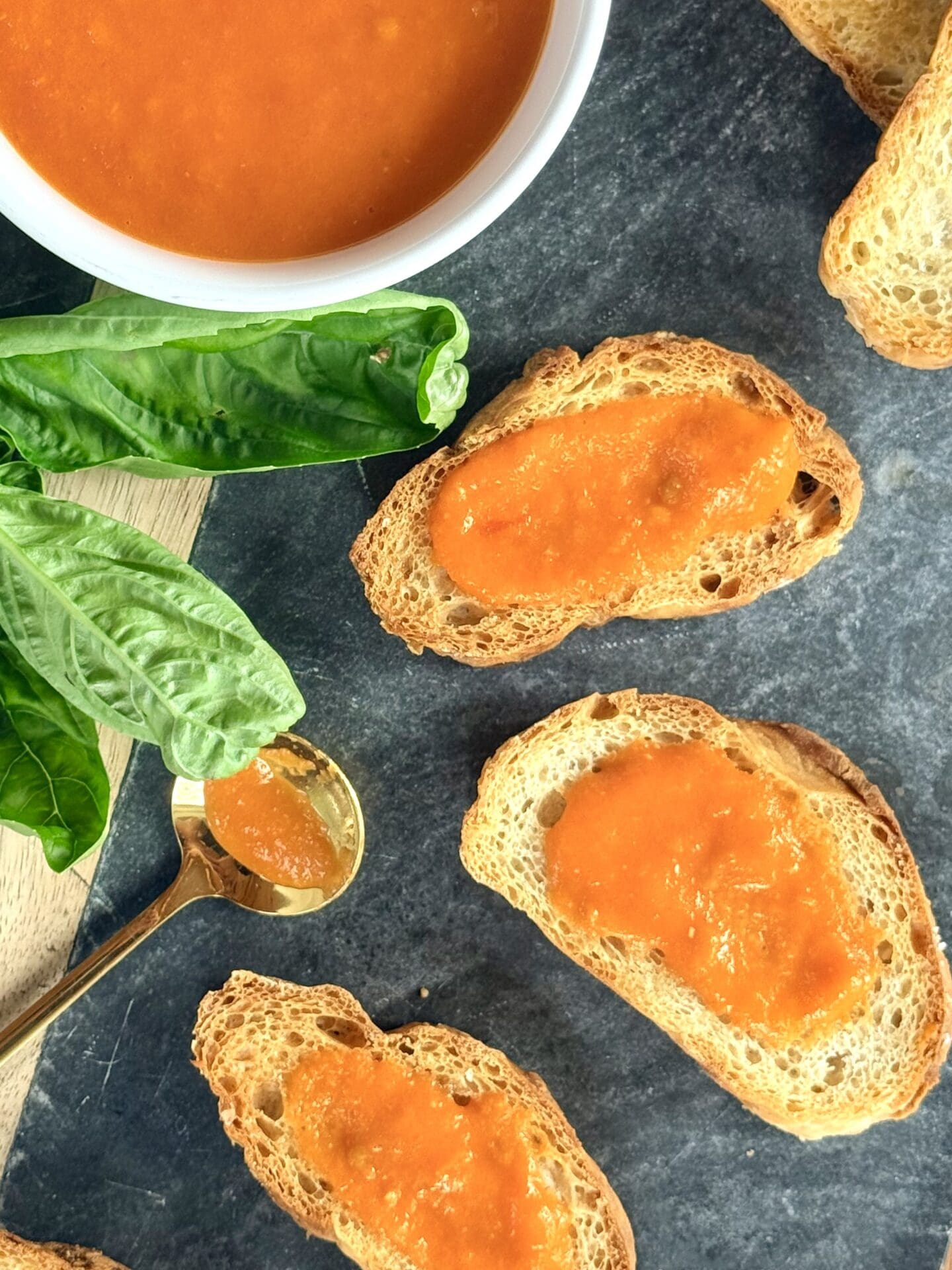 Crostini topped with fresh tomato sauce are seen from above. A bowl of the sauce and a sprig of fresh basil sit nearby.