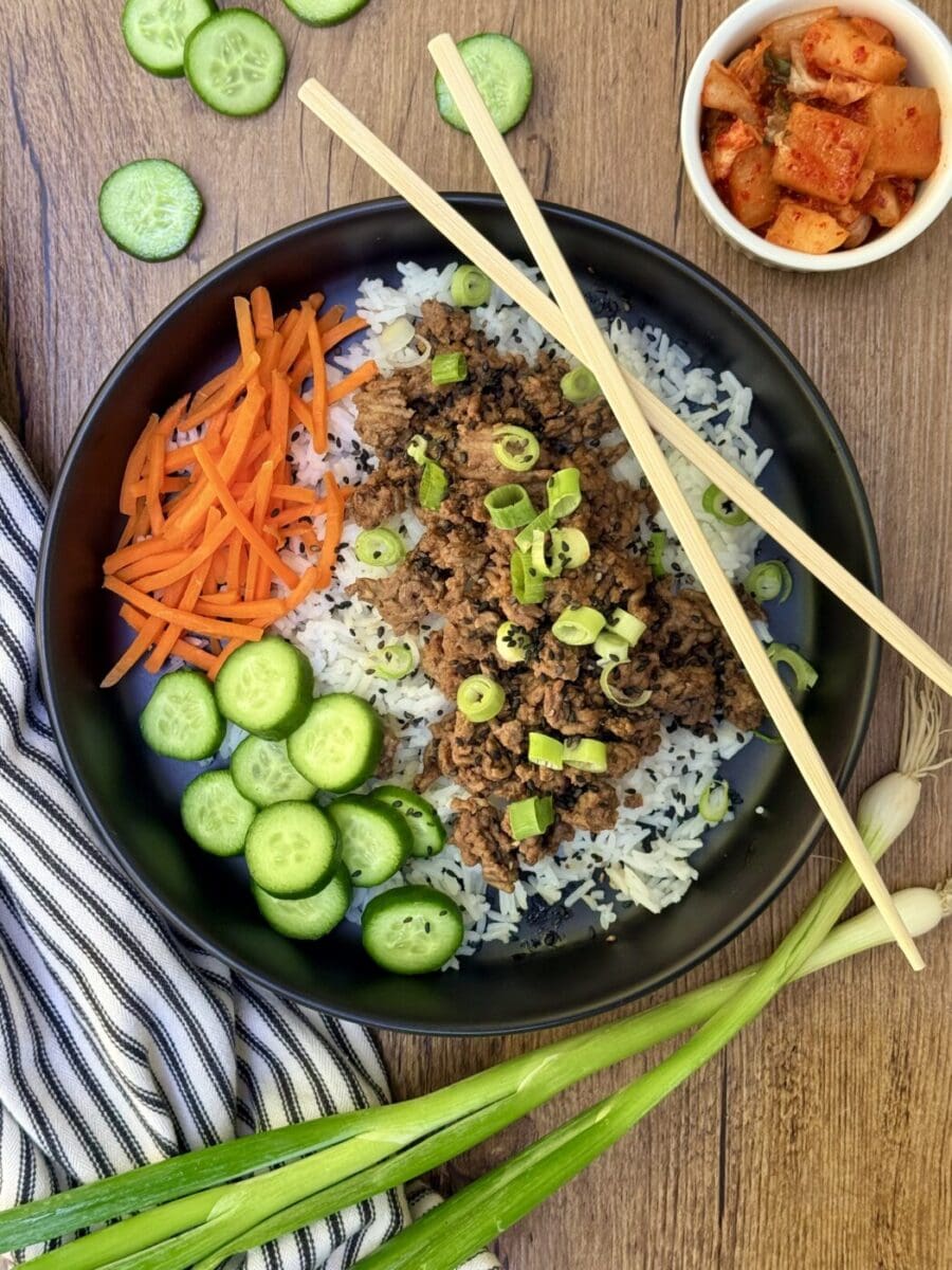 A black bowl filled with Korean Ground Beef, vegetables and rice is seen from above, along with a set of chopsticks. A dish of kimchi sits nearby.