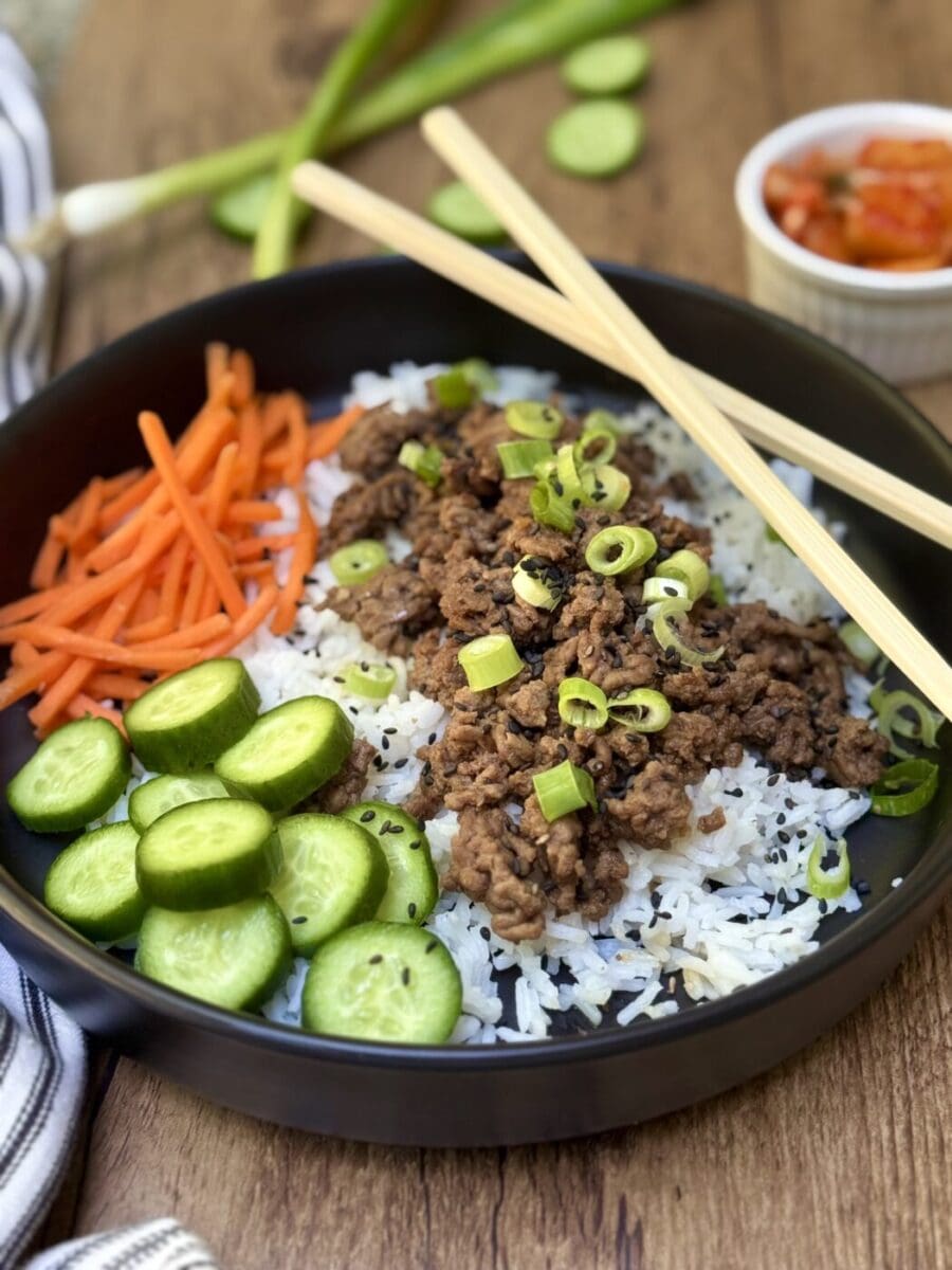 Korean Ground Beef Bowl on a wood table. A Korean Ground Beef Bowl is seen on a wood table, surrounded by fresh vegetables, chopsticks and kimchi.