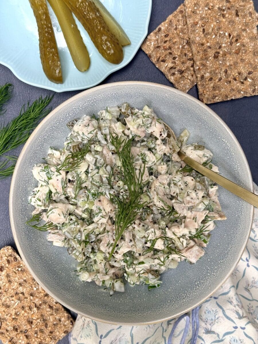 A bowl of Greek Yogurt Dill Pickle Chicken Salad is seen from above, surrounded by crisp bread, fresh dill and sliced pickles.