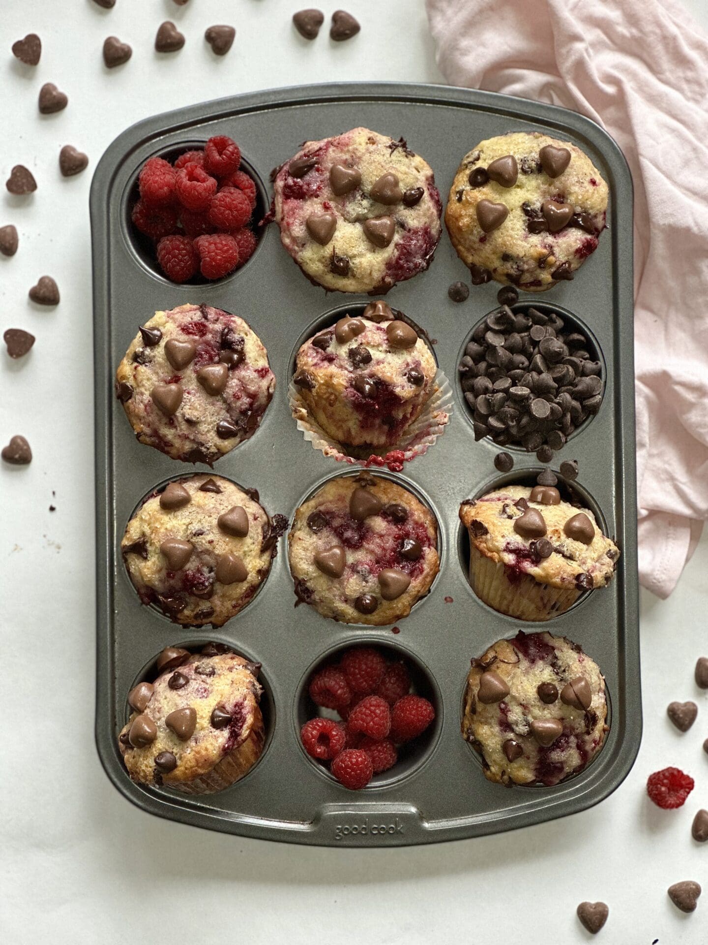 Muffin pan full of decadent raspberry chocolate muffins surrounded by fresh raspberries and heart shaped chocolates