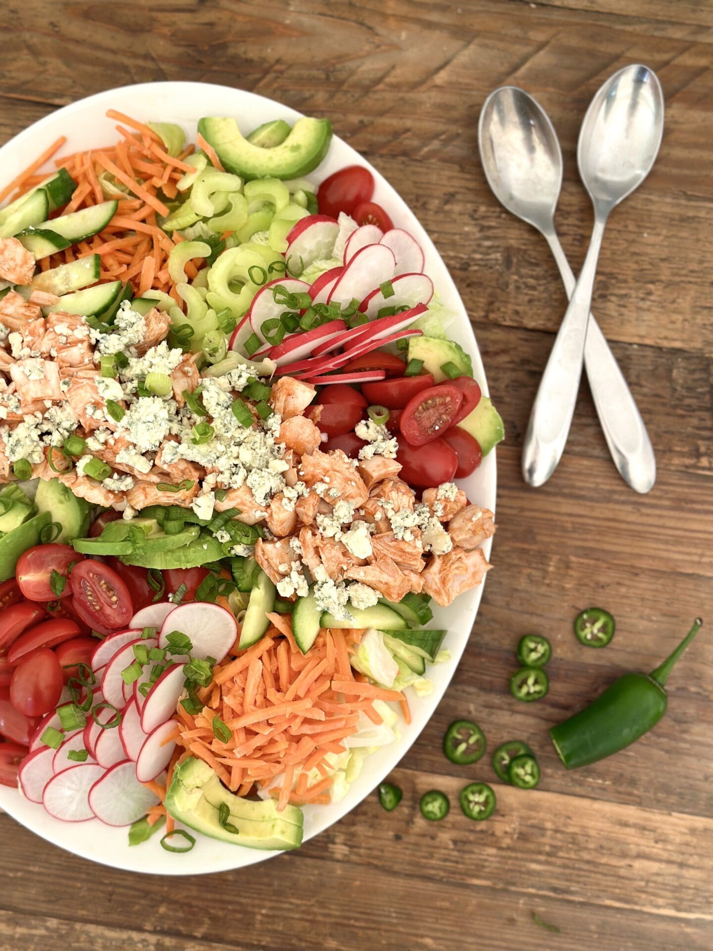 IMG_9349 A large platter of Buffalo Chicken Dinner Salad full of bright and crisp vegetables is seen from above on a wood table with serving spoons and sliced jalapeños
