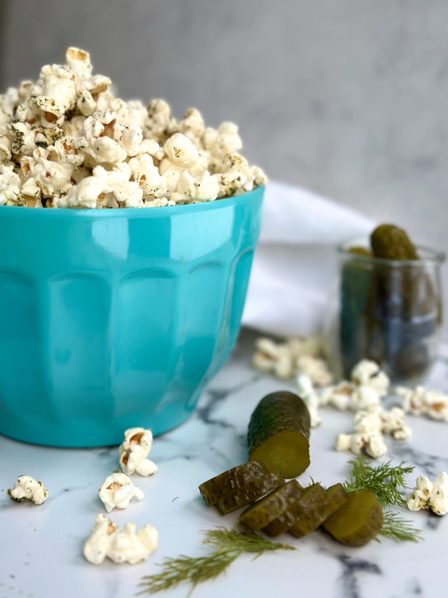 IMG_0952 A turquoise bowl of Dill Pickle popcorn is seen on a white marble table surrounded by green dill pickles, fresh dill sprigs and popcorn pieces