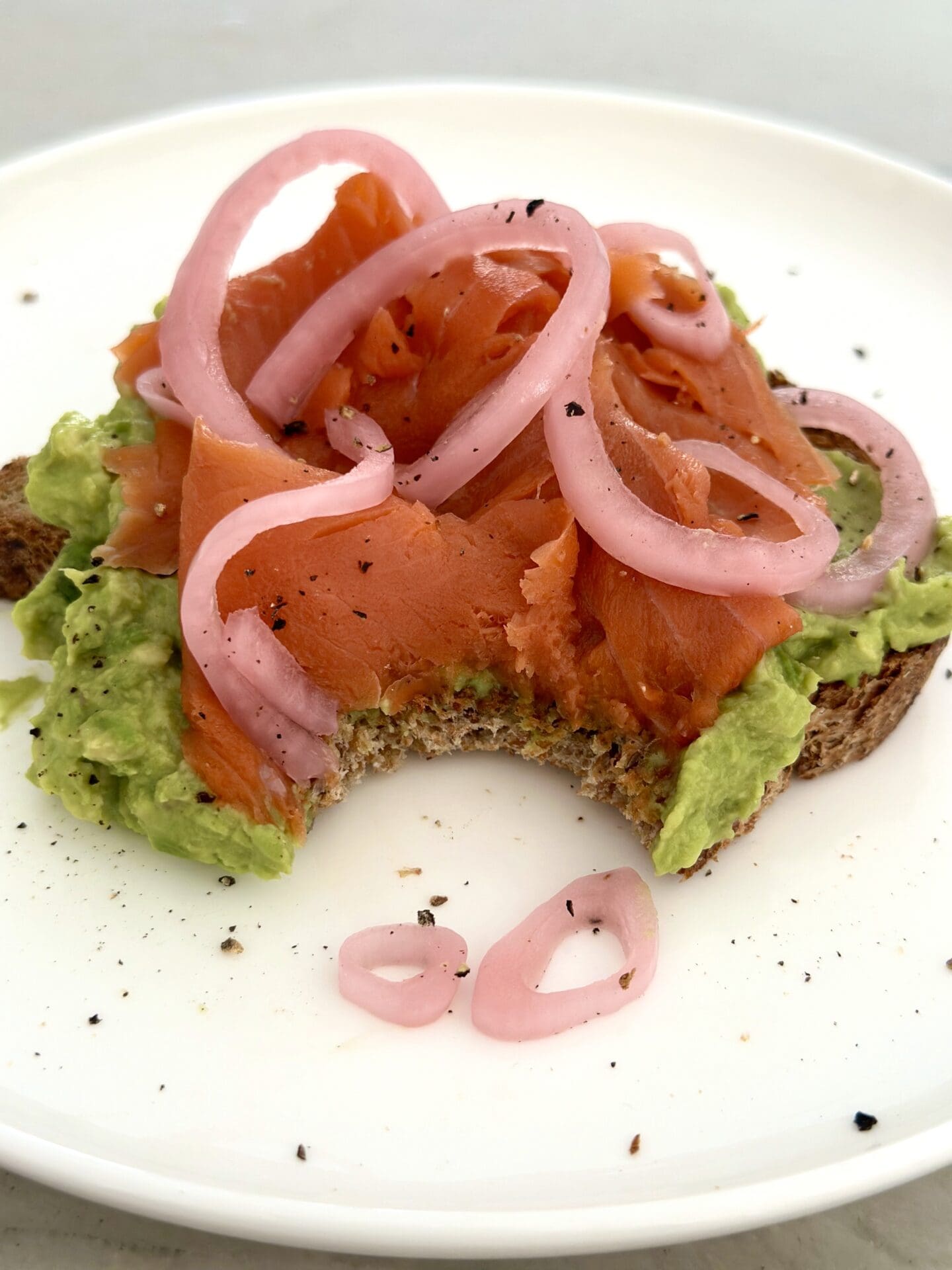 Avocado toast with smoked salmon and pickled red onions sits on a white plate. A large bite has been taken out of the avocado toast