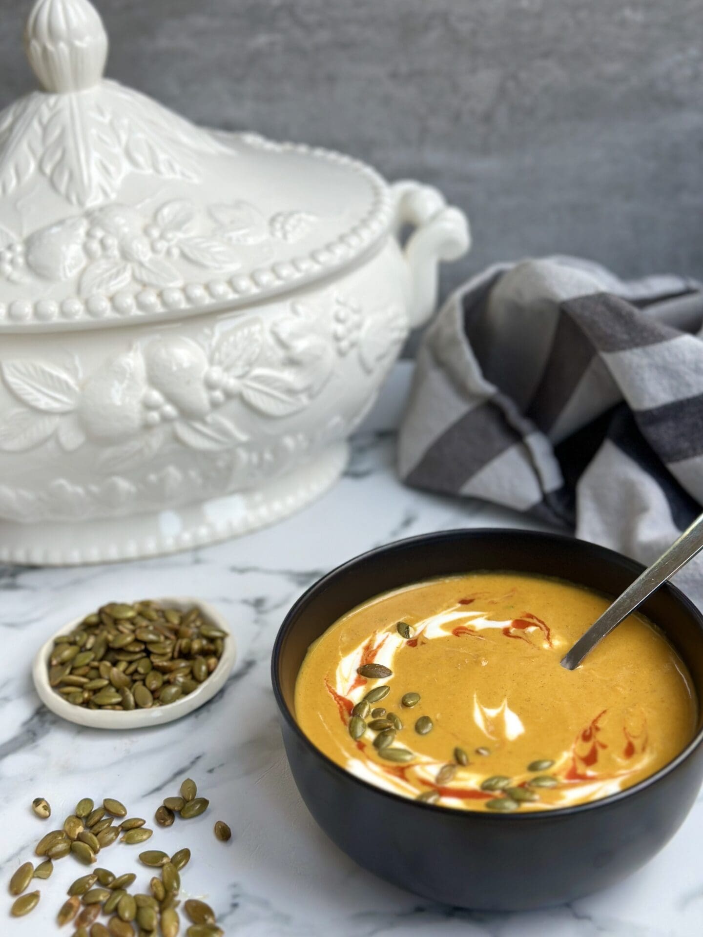 IMG_9788 A black bowl of golden Creamy Pumpkin Spice Soup is seen on a white marble table surrounded by a a soup tureen and a small bowl of pumpkin seeds