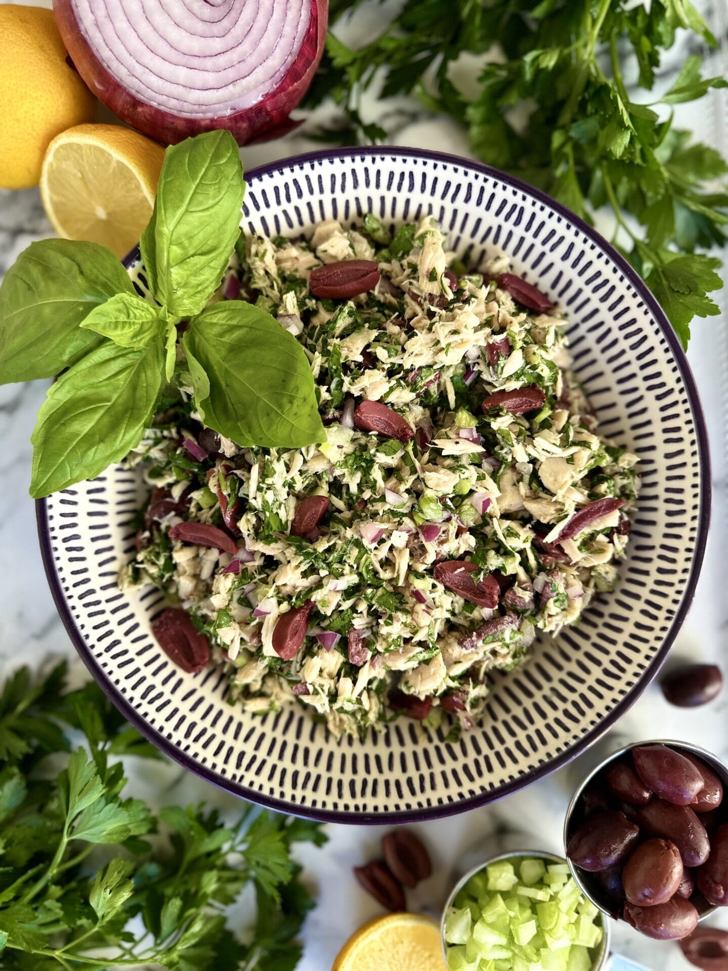 A bowl of Italian tuna salad is seen from above, garnished with fresh basil leaves. The bowl sits on a white marble table and is surrounded by parsley, lemons, red onions and black olives.