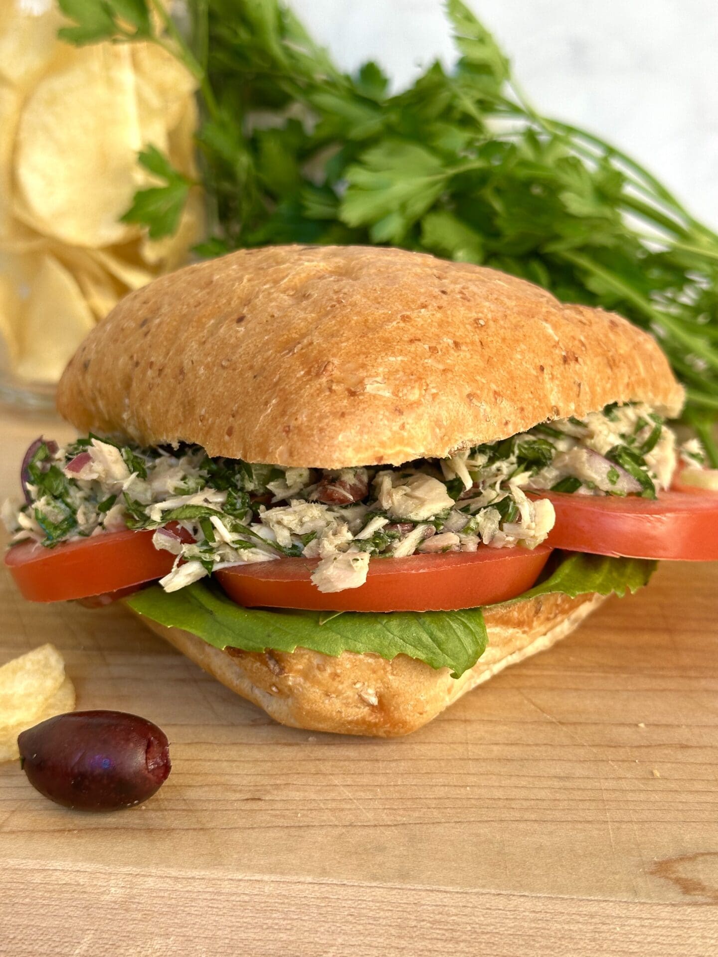 A large ciabatta bun is stuffed full of Italian tuna salad, thick red tomato slices and fresh basil leaves. The sandwich sits on a wood serving board, surrounded by black olives, potato chips and parsley.