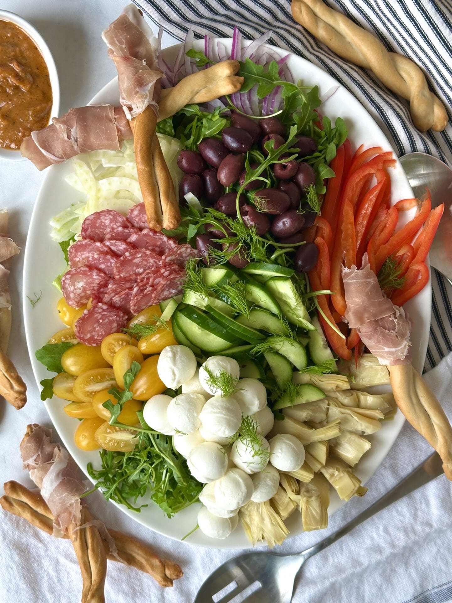 A platter of Antipasti Salad with Prosciutto Wrapped Bread Sticks is seen from above. The platter overflows with fresh, colourful vegetables, olives, salami and cheese and is surrounded by a bowl of sundried tomato dressing and prosciutto wrapped breadsticks.