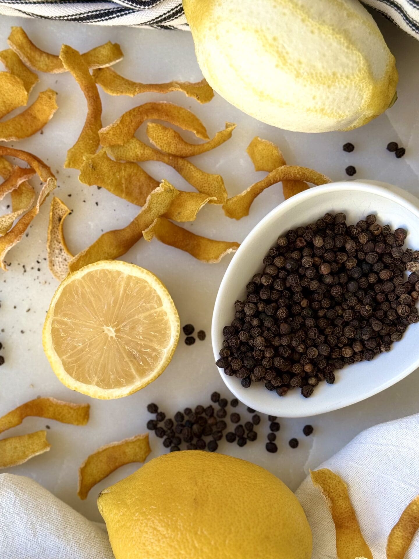 IMG_4120 The ingredients for Homemade Lemon Pepper Seasoning - lemon zest, lemons and peppercorns - are seen from above, on a white marble table.