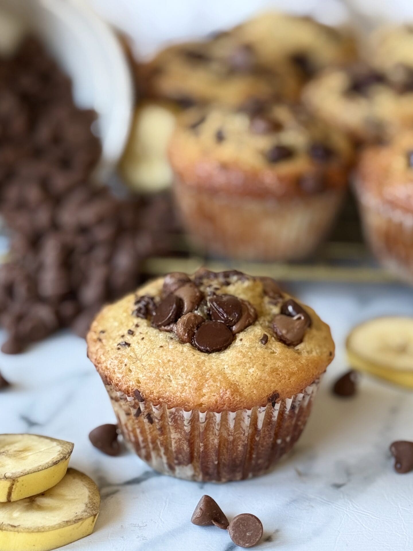 A single Sour Cream Banana Chocolate Chip Muffin is seen in the foreground, topped with loads of chocolate chips. In the background, more muffins rest on a cooling rack beside a bowl of chocolate chips.