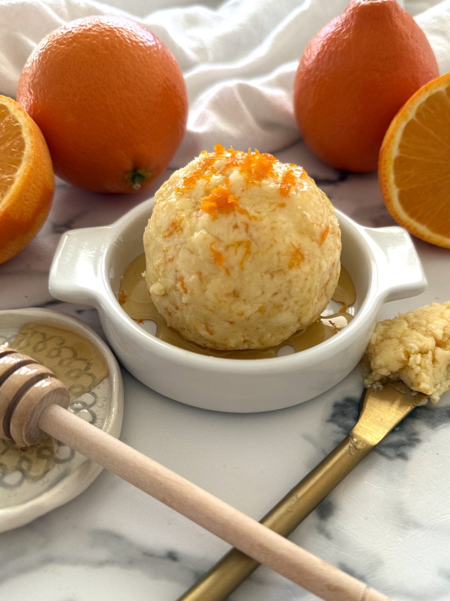Orange and Honey Butter is shaped into a ball, placed in a shallow butter dish, drizzled with extra honey and garnished with fresh orange zest. Fresh oranges and a honey dipper complete the image.