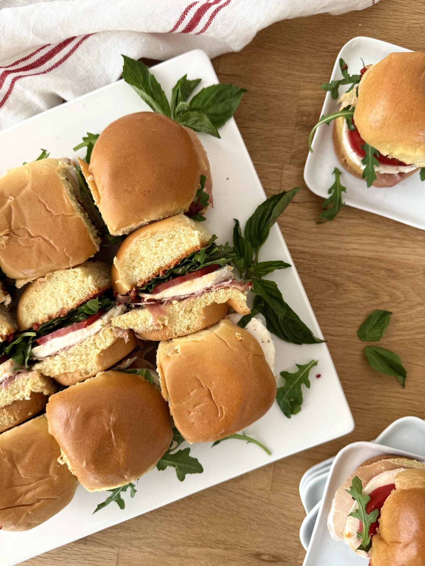 A platter of Italian Antipasto Sliders is seen from above, surrounded by individual plates of sandwiches and fresh basil leaves.