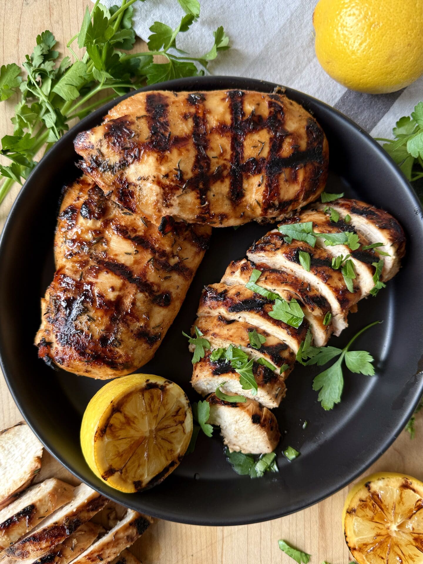 IMG_5156 A black platter is seen from above, filled with Sweet and Tangy Marinated Grilled Chicken. One chicken breast is sliced, whereas the others are seen whole, with perfect crosshatch grill marks. The chicken is garnished with grilled lemon halves and fresh parsley.