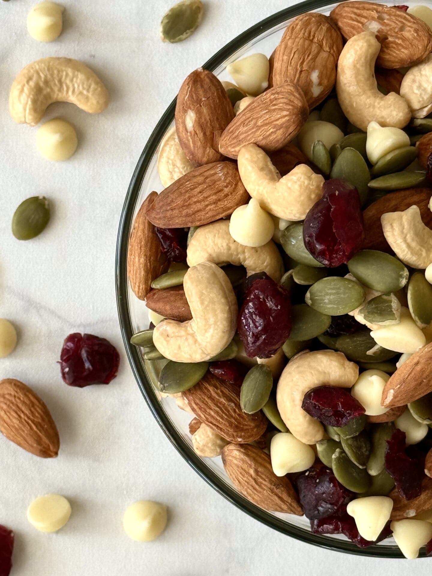 A glass bowl of White Chocolate and Cranberry Trail Mix is seen from above. Cashews, pumpkin seeds, dried cranberries and white chocolate chips are seen scattered around the bowl.