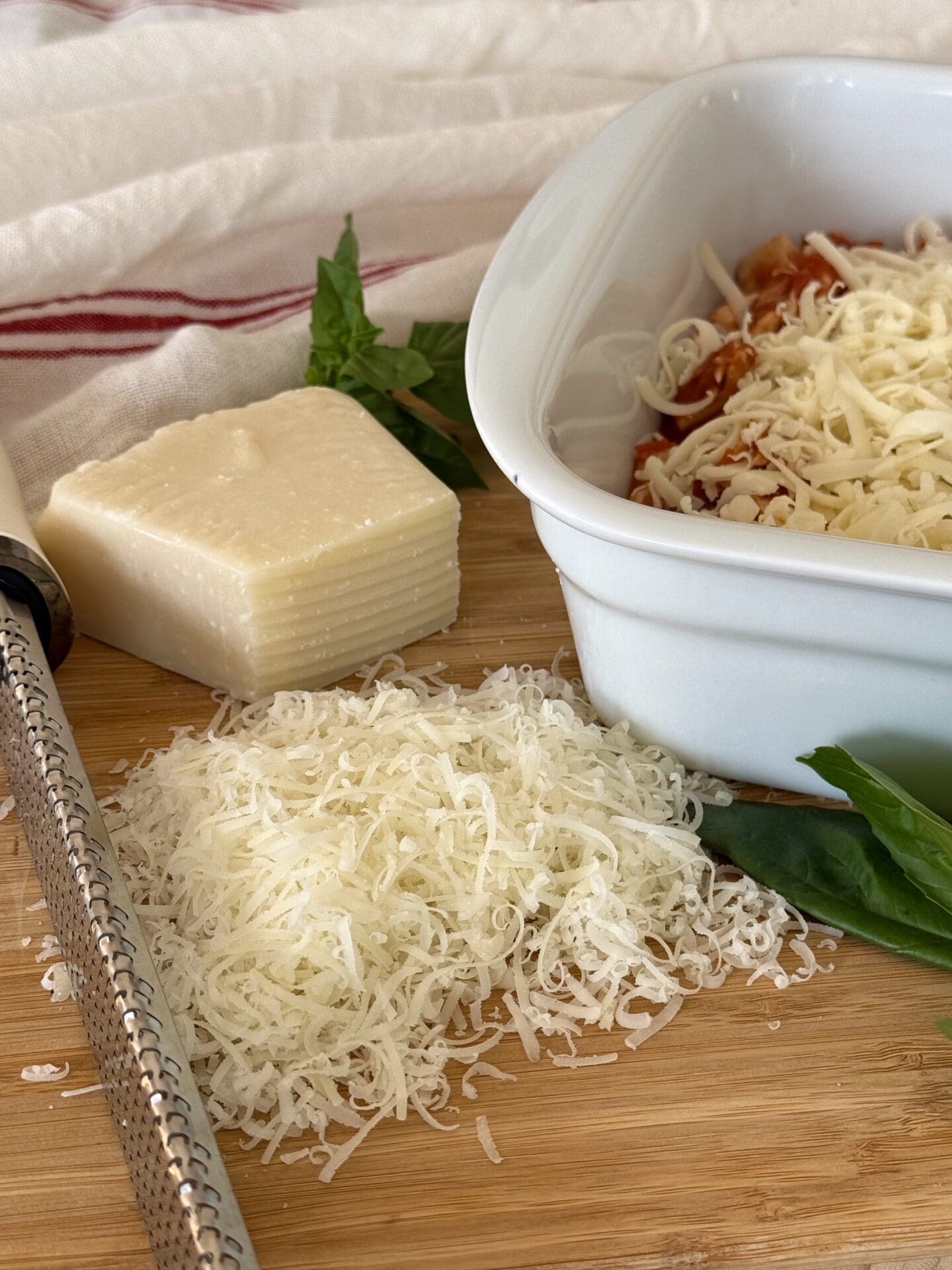 Healthier Chicken Parmesan Casserole is seen being assembled. A white baking dish filled with marinara sauce, chopped cooked chicken and mozzarella cheese sits off to the side. A wedge of fresh parmesan cheese is being grated with a rasp.