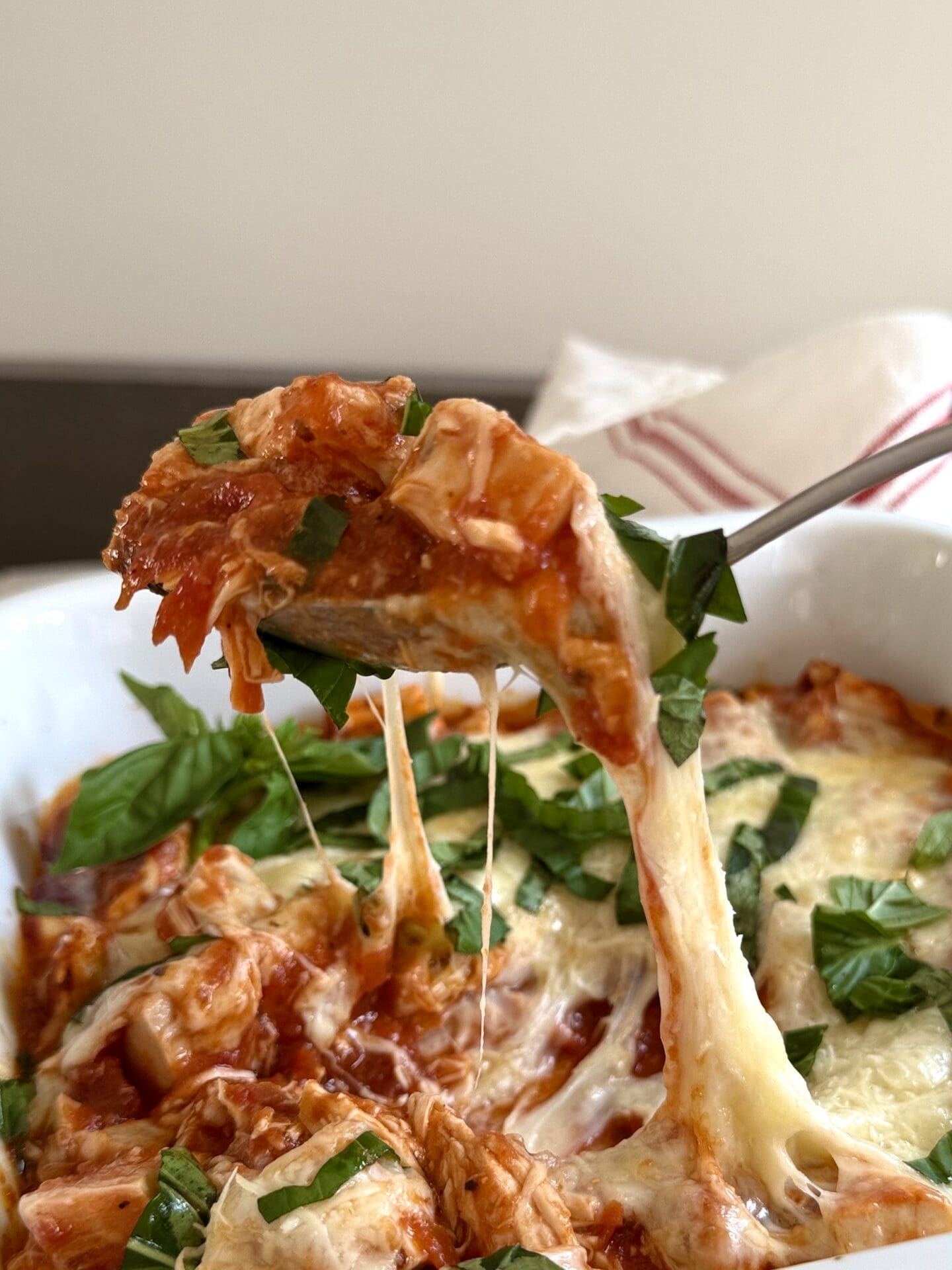A large spoon scoops up a hearty portion of Healthier Chicken Parmesan. Melty mozzarella cheese stretches from the casserole to the spoon.