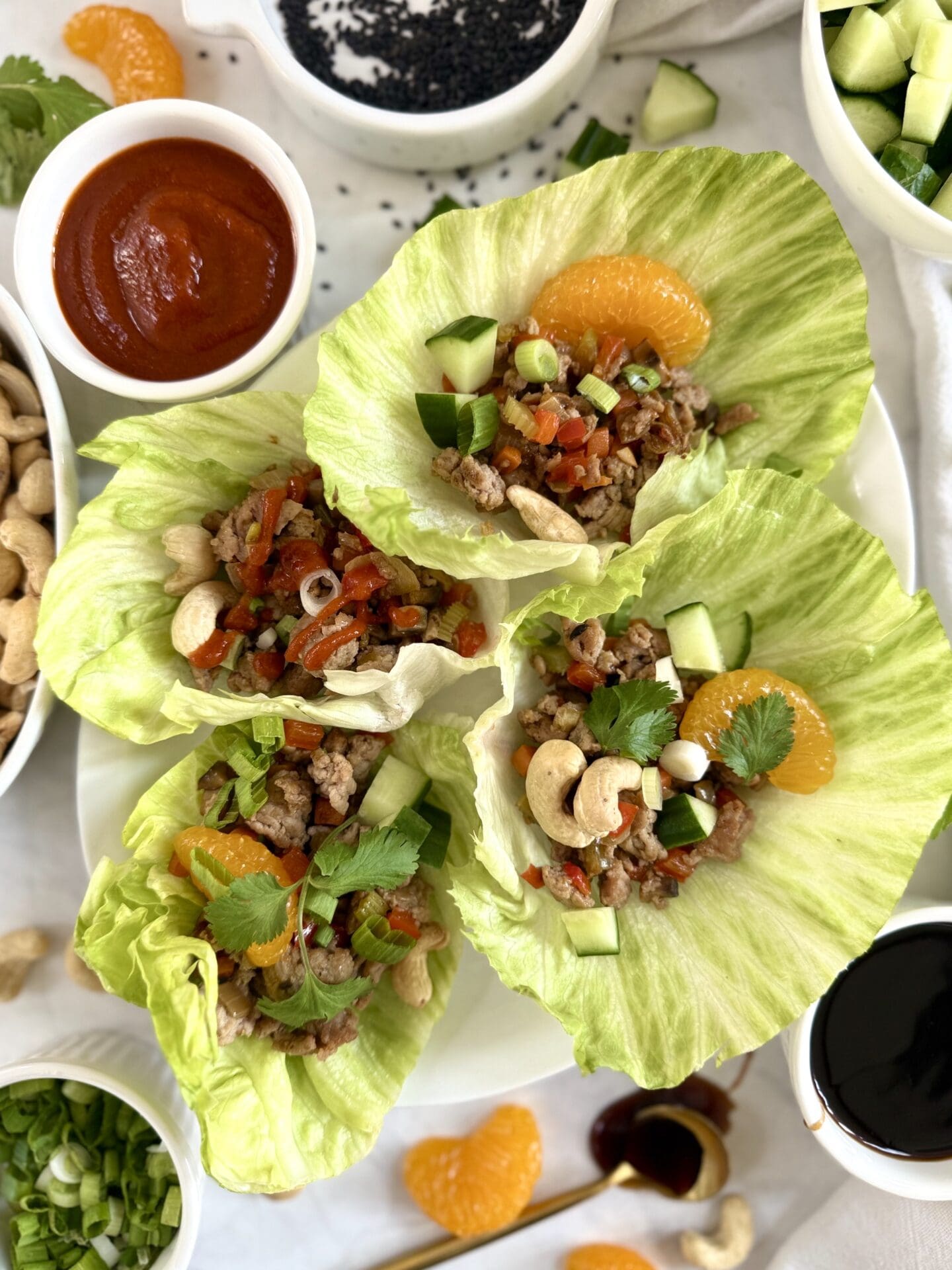 A plate of Ground Turkey Lettuce Wraps is seen from above, surrounded by dishes of chopped cucumber, spring onions, hoisin sauce, cashews and sriracha sauce.