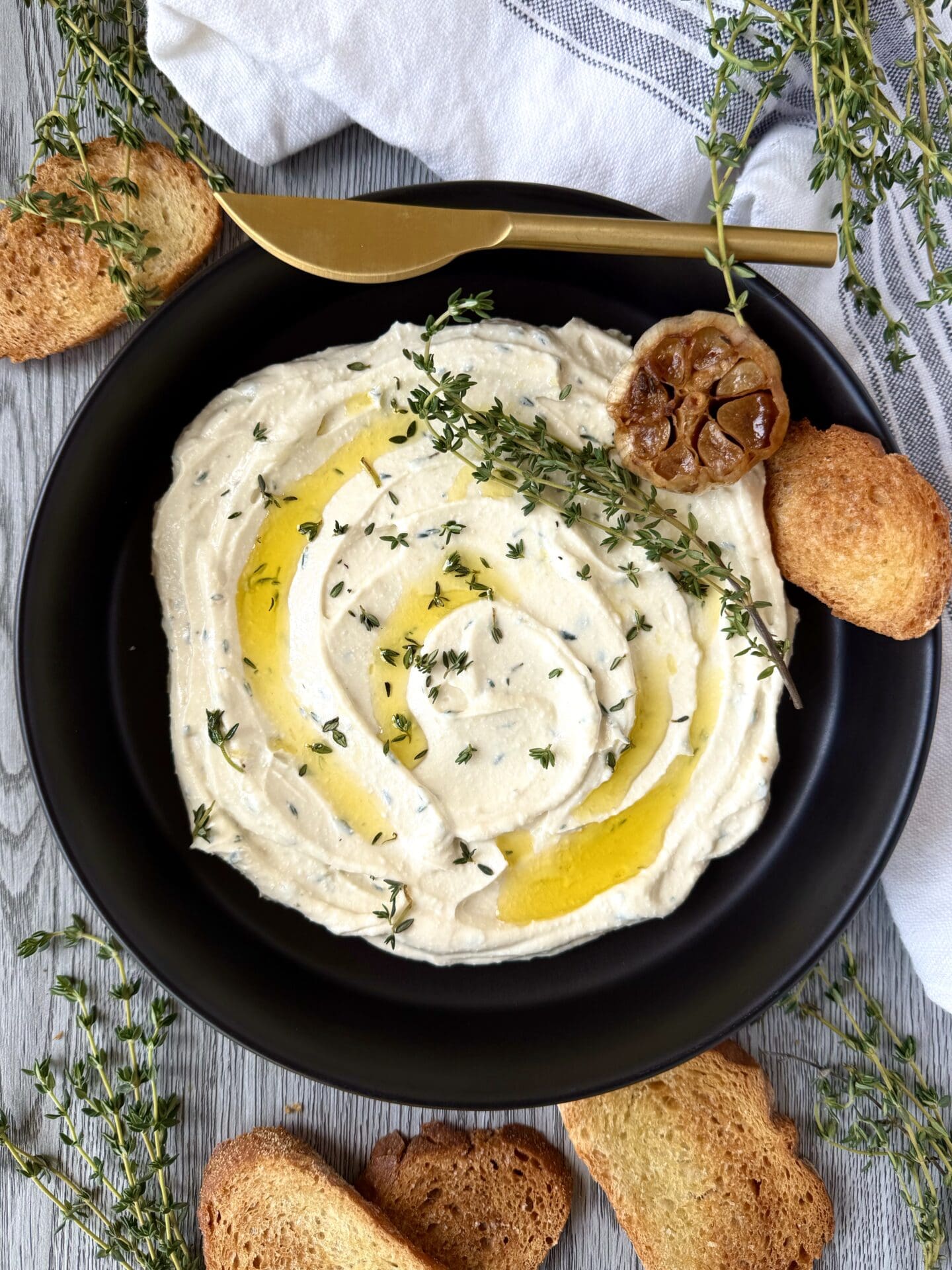 A black bowl of Whipped Feta with Roasted Garlic and Fresh Thyme is seen from above, garnished with olive oil, fresh thyme and a head of roasted garlic. Sprigs of fresh thyme and slices of toasted bread surround the bowl.