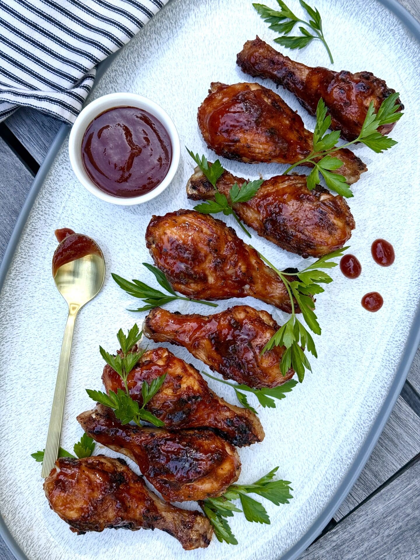 A platter containing Smokey Sweet Grilled Chicken Legs lined up in a row is seen from above. A dish of barbecue sauce with a dripping spoon rests on the side. The platter is garnished with plenty of vibrant, fresh parsley.