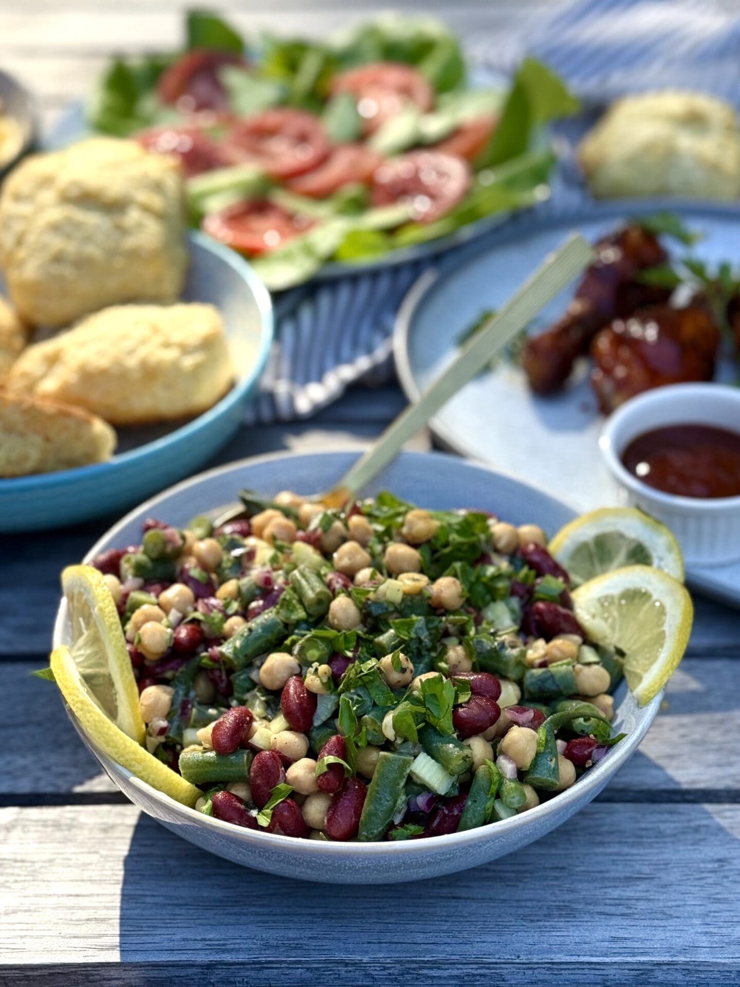 A bowl of Marinated Mediterranean Bean Salad sits in the late afternoon light on a picnic table surrounded by platters of grilled chicken, biscuits and salads.