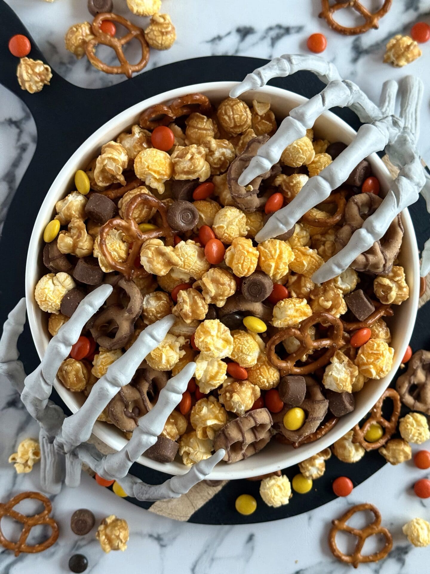 A bowl of Monster Munch Halloween Snack Mix is seen from above. Skeletons hands reach into the bowl from either side.
