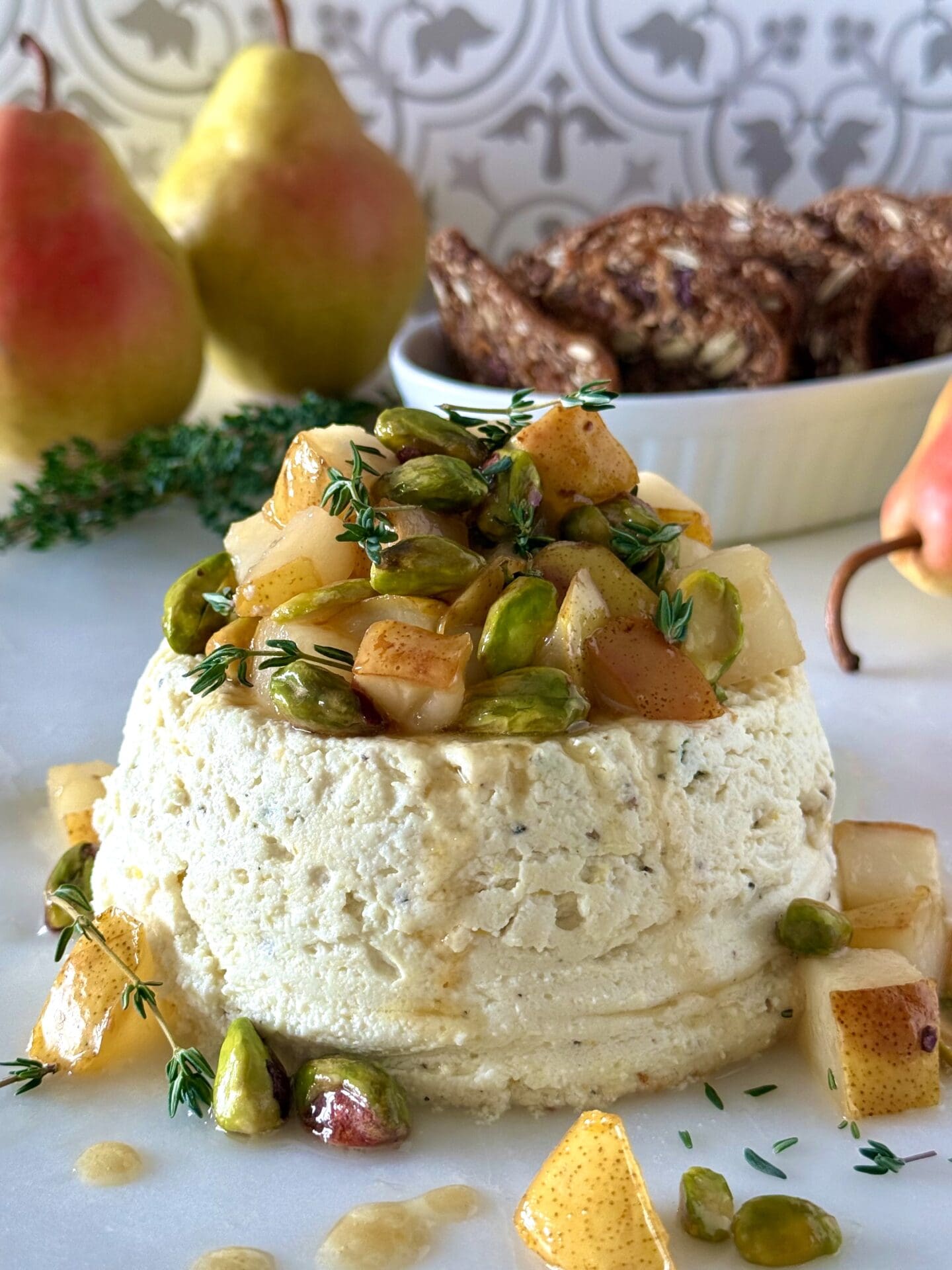 An elegant Pistachio and Pear Baked Ricotta sits in the foreground with chunks of caramelized pear, bright green pistachio and fresh sprigs of thyme tumbling from the top. The appetizer is surrounded by fresh pears and crackers.