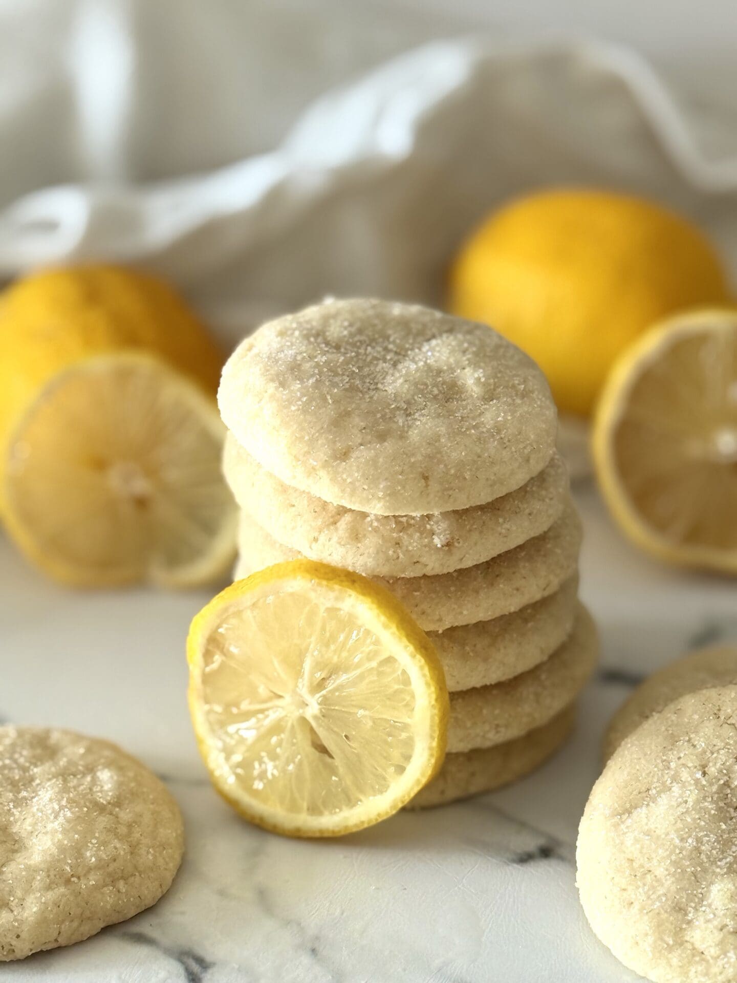IMG_7401 A stack of glittering chewy lemon sugar cookies is seen on a white marble countertop, surrounded by whole lemons and fresh lemon slices.