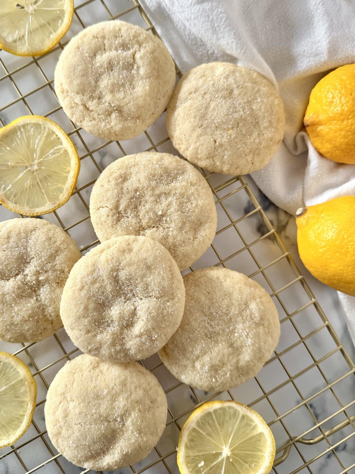IMG_7410 Freshly baked soft and chewy lemon sugar cookies are seen from above, cooking on a gold wire rack, surrounded by lemon slices.
