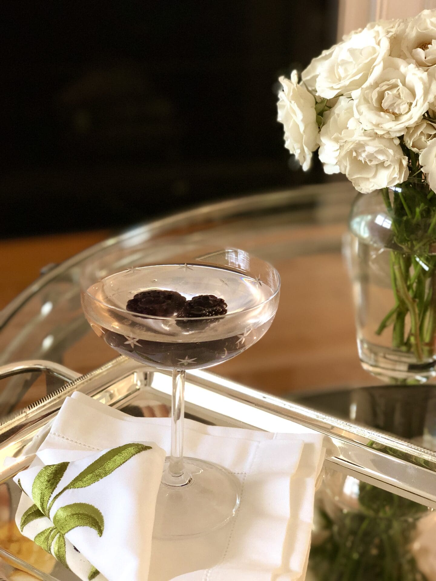 IMG_0662 A pink Sparkling Blackberry Blush cocktail is seen in a coupe glass on a silver platter. Fresh blackberries float in the drink. A vase of white roses is seen in the background.