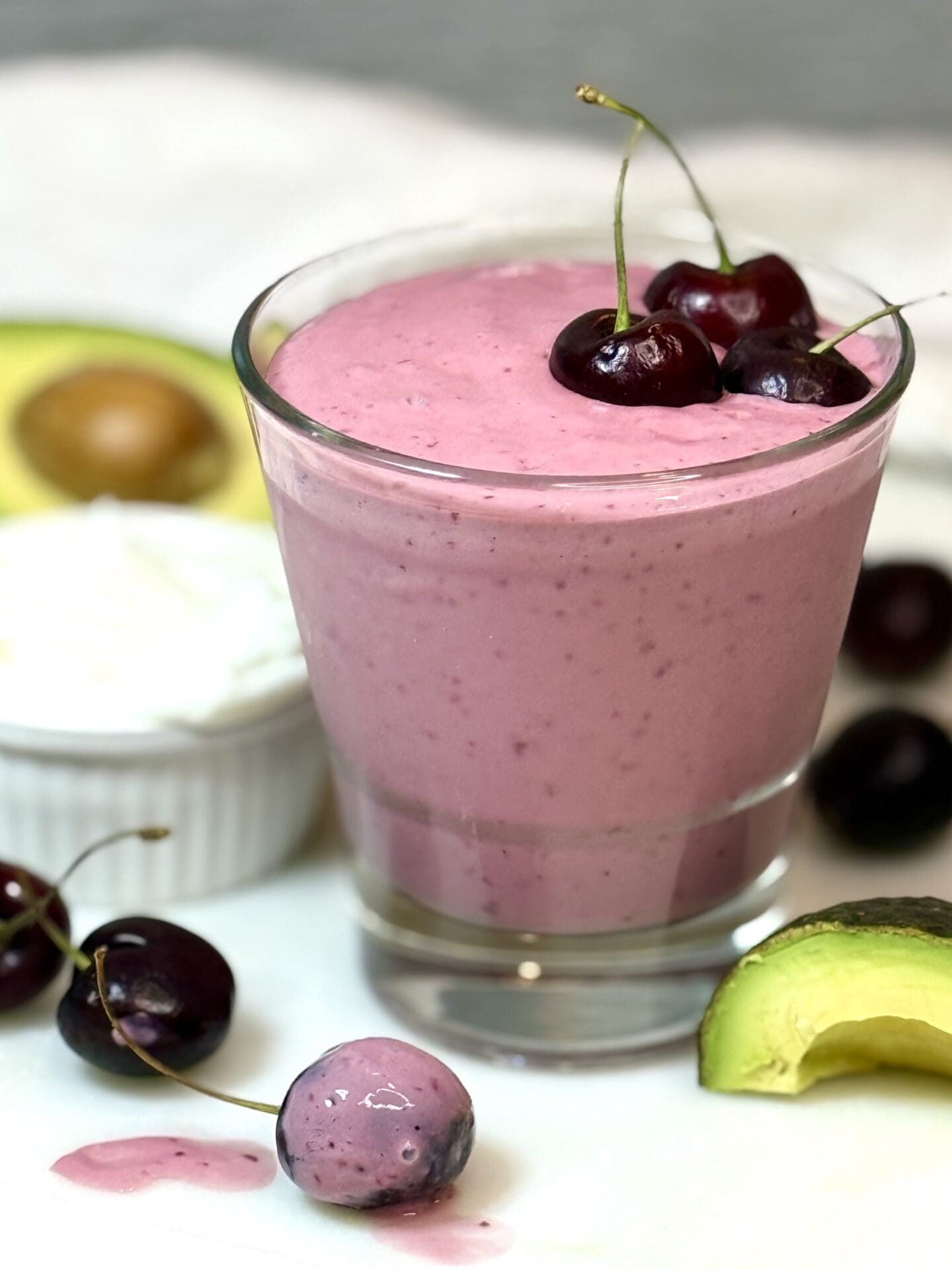 A glass of pink cherry vanilla smoothie is seen on a white marble counter. The glass is garnished with fresh cherries and is surrounded by fresh avocado and more cherries.