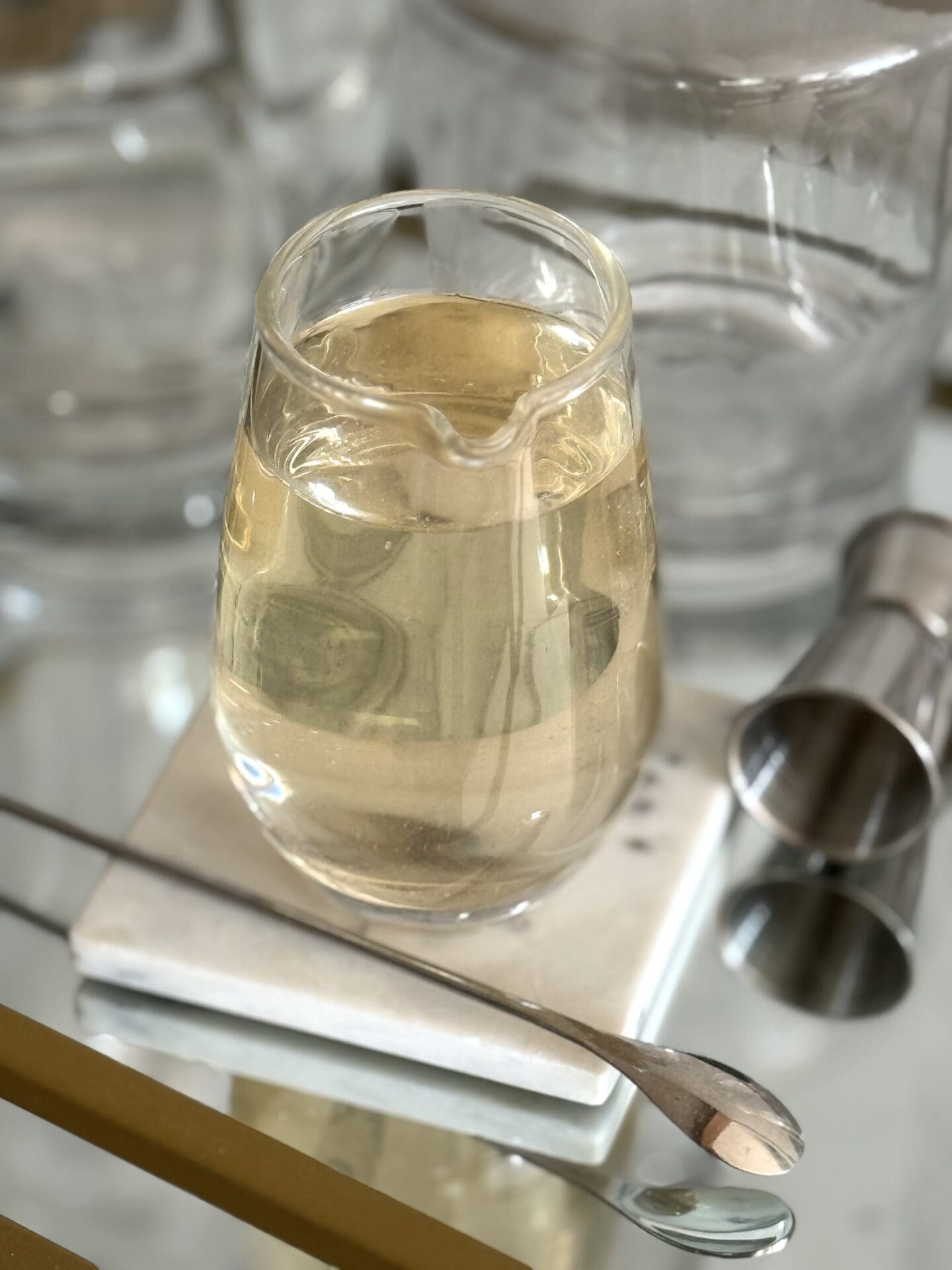 A glass of homemade simple syrup sits on a sparkling bar cart, surrounded by a jigger, stirring spoon and other bar implements.