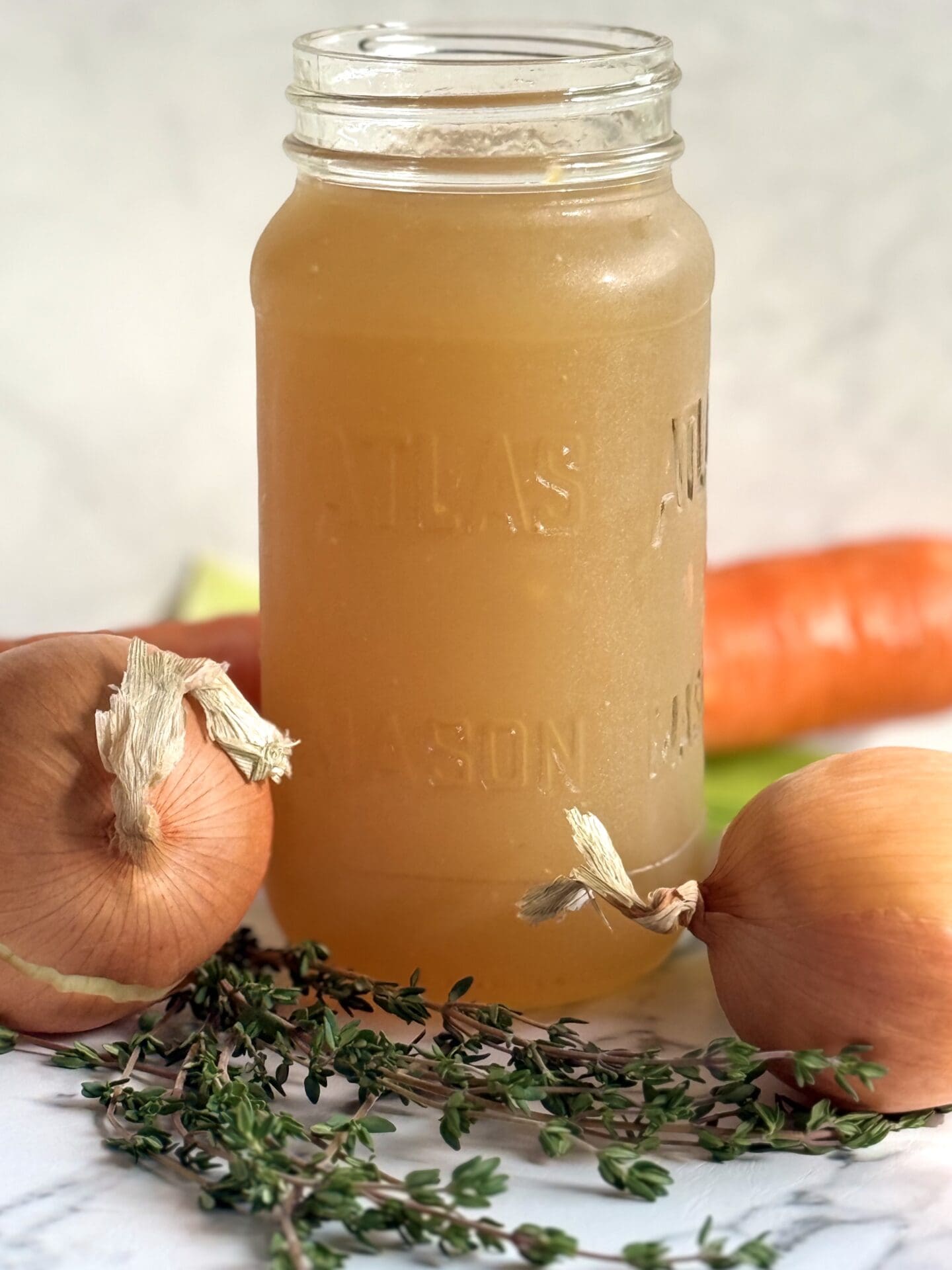 IMG_8147 A glass jar of homemade chicken stock sits on a marble counter, surrounded by carrots, celery, onions and fresh thyme.