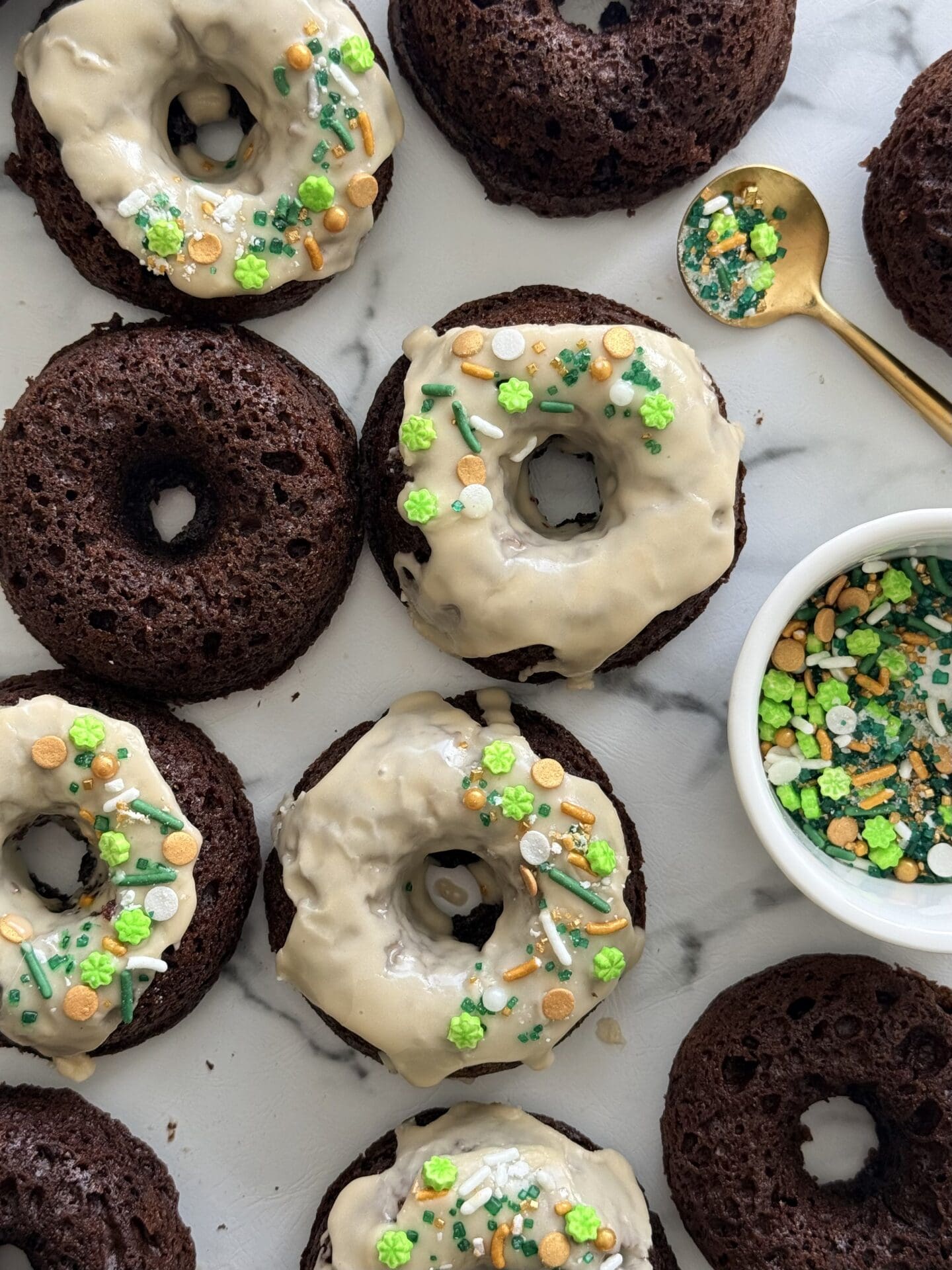 A group of baked chocolate donuts is seen from above. Some are topped with a Baileys glaze and St Patrick’s Day themed sprinkles.