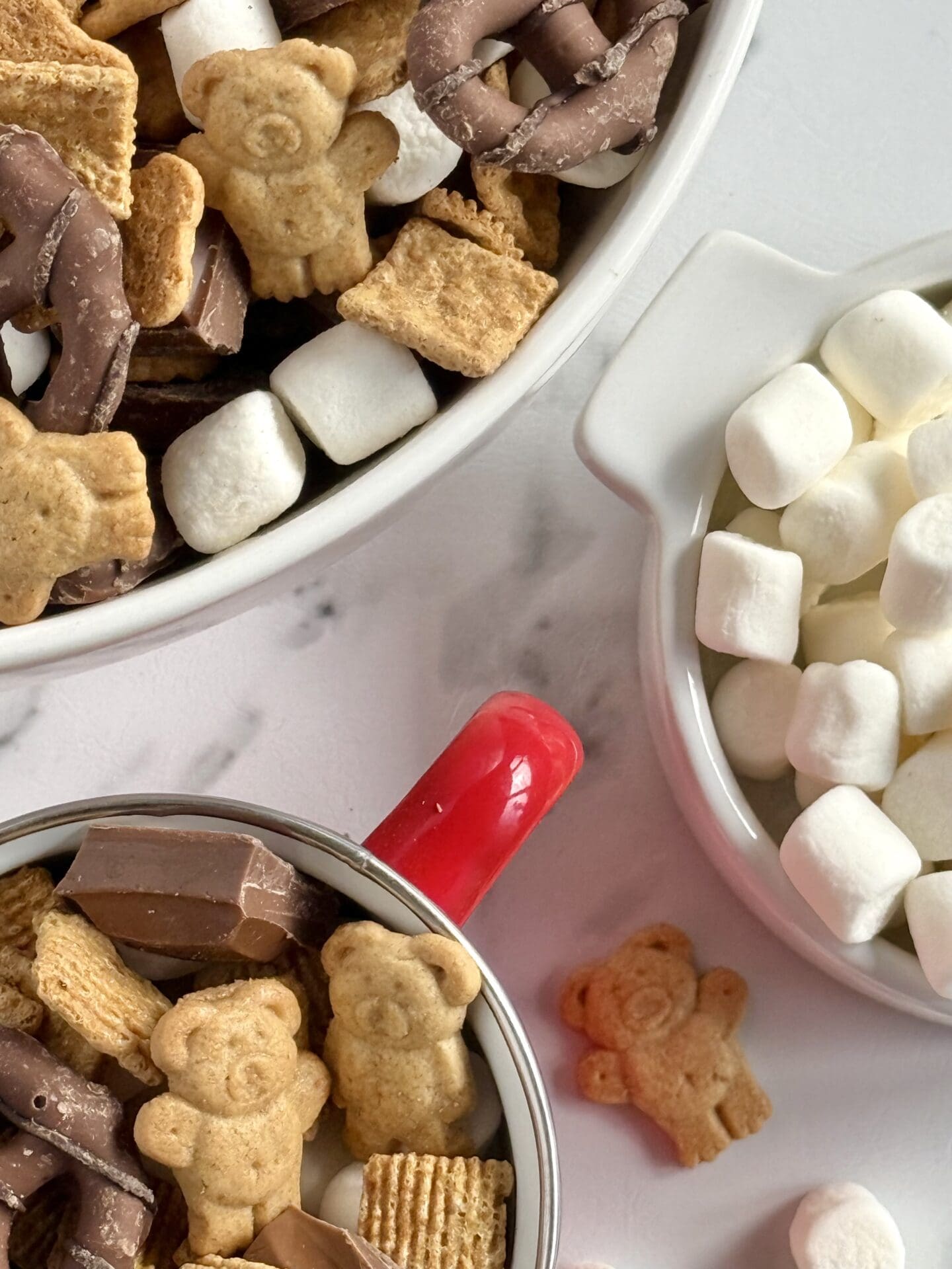 IMG_7055 Three bowls of S’mores Snack Mix and mini marshmallows are seen from above. Marshmallows and Teddy Grahams spill from the bowls onto the white marble counter.