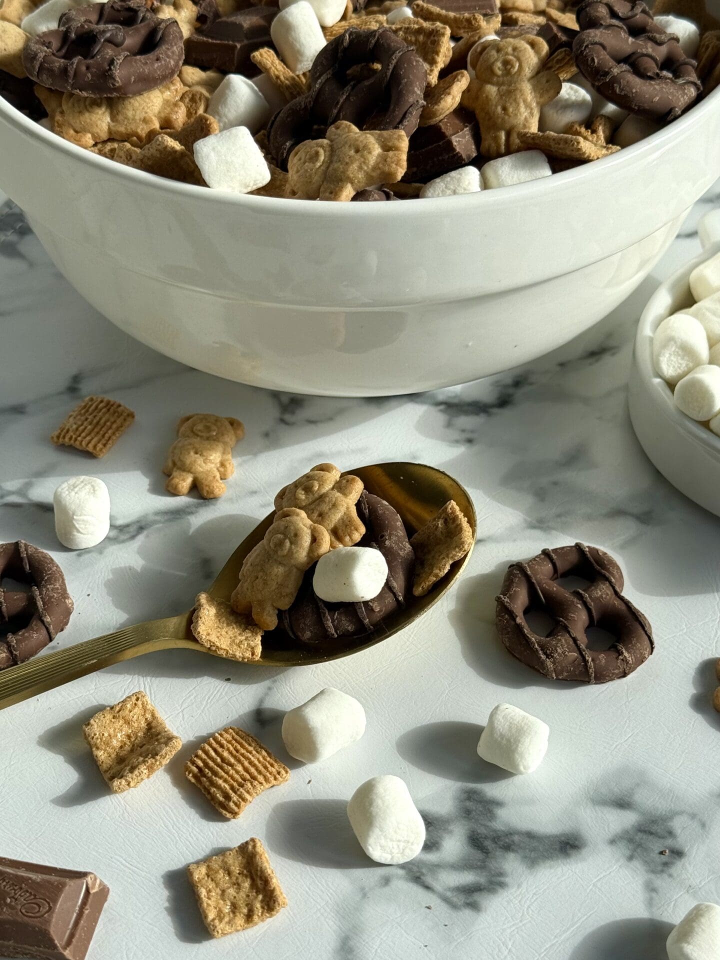 IMG_7063 A large bowl of S’Mores Snack Mix sits on in the sunlight. A gold spoon appears in the foreground, spilling chocolate covered pretzels, marshmallows, Golden Grahams and chunks of milk chocolate onto the counter.