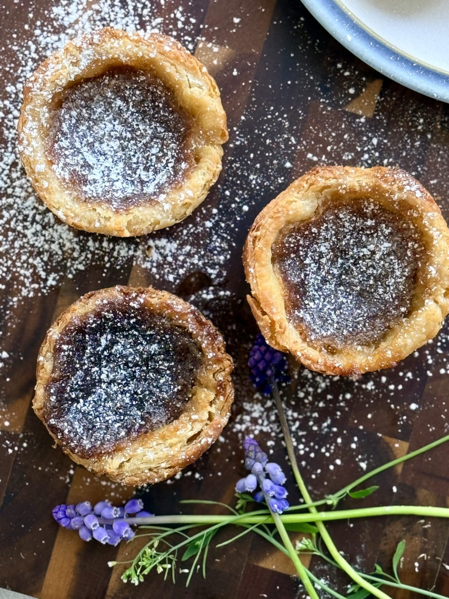 IMG_8452 The Lakehouse Butter Tarts are seen from above, dusted in powdered sugar and resting on a wooden board. Small purple and white flowers complete the image.